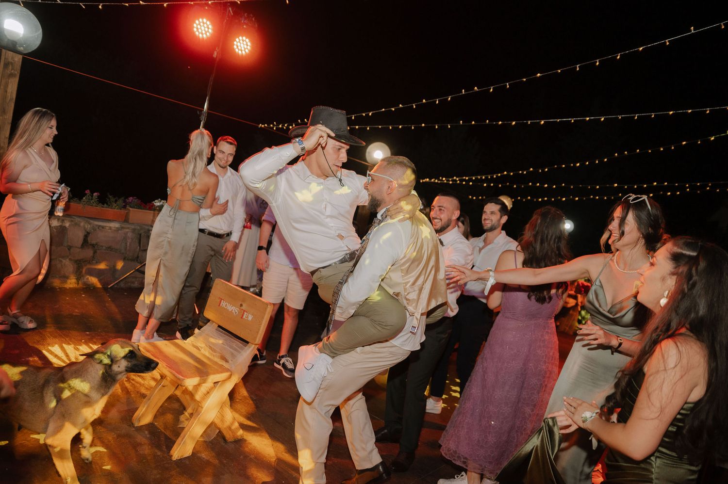 People dancing and celebrating at an outdoor evening party with string lights overhead.