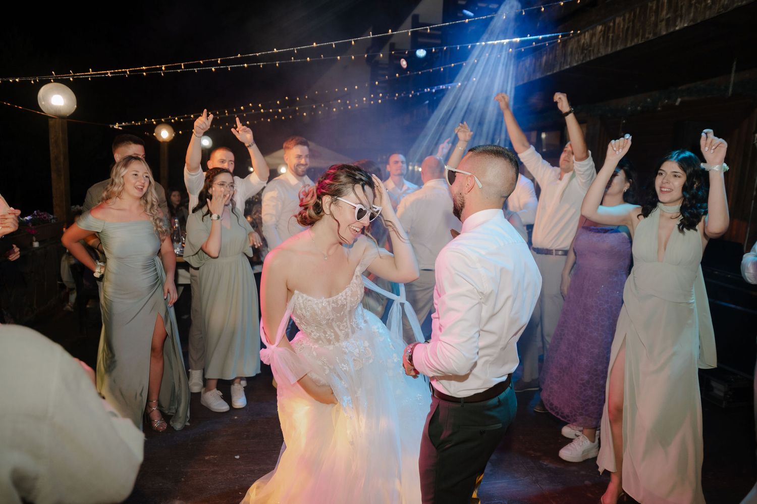Wedding party dancing in a circle under blue spotlights and string lights during an evening outdoor celebration.