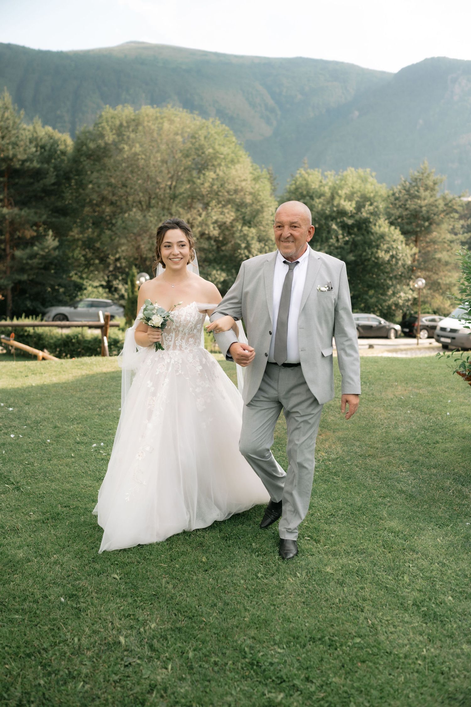 A couple in wedding attire runs playfully through a green field with mountains in the background on their special day.