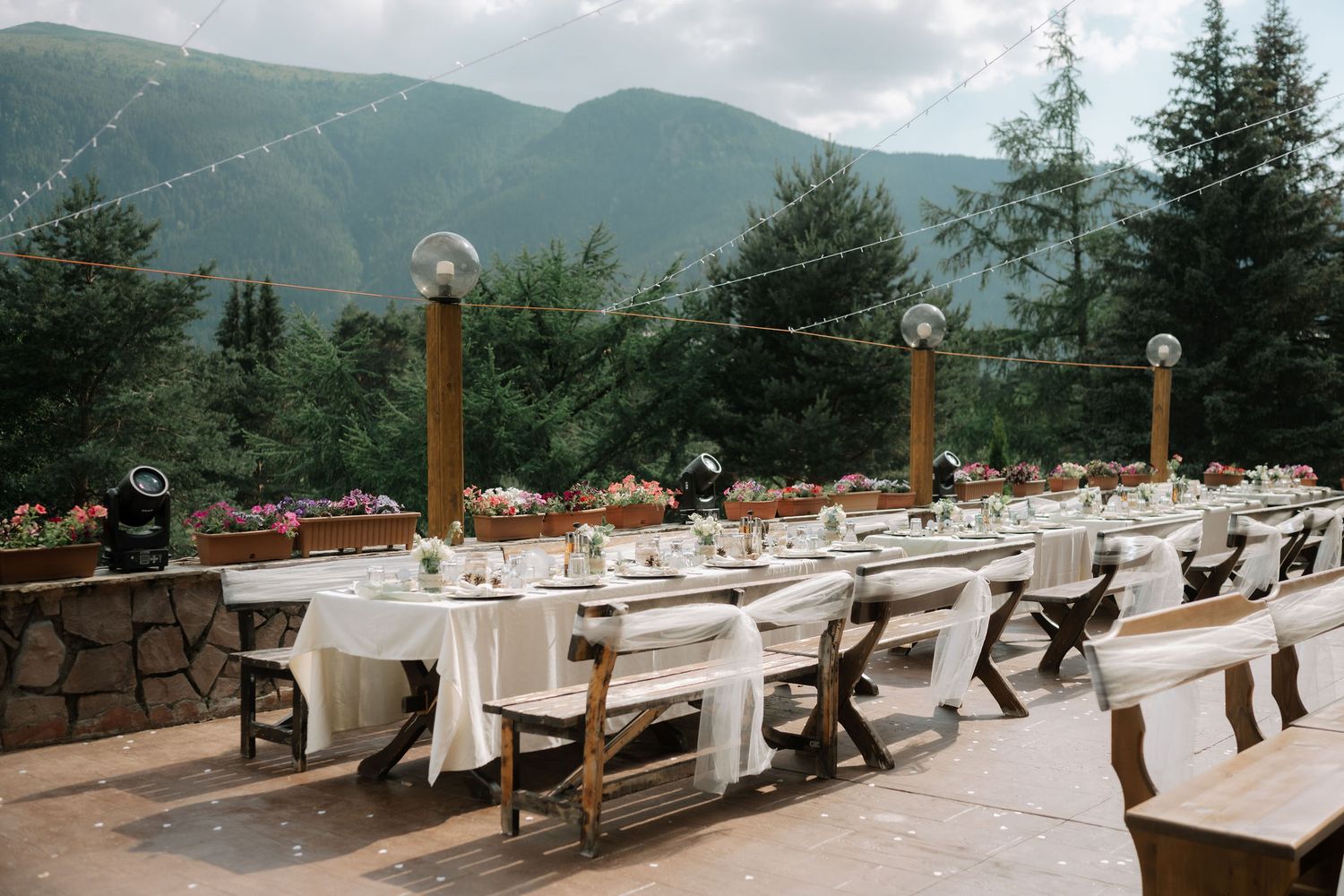 Elegant outdoor wedding reception setup with long tables and benches on a stone patio overlooking misty mountain views.
