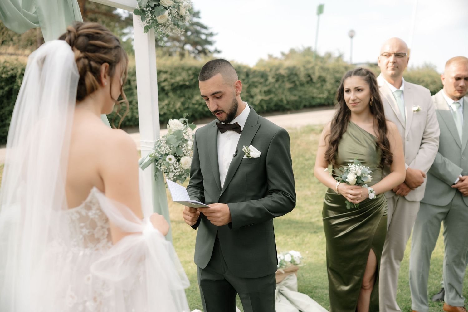 A wedding ceremony under a white arch with guests in formal attire gathered outdoors.