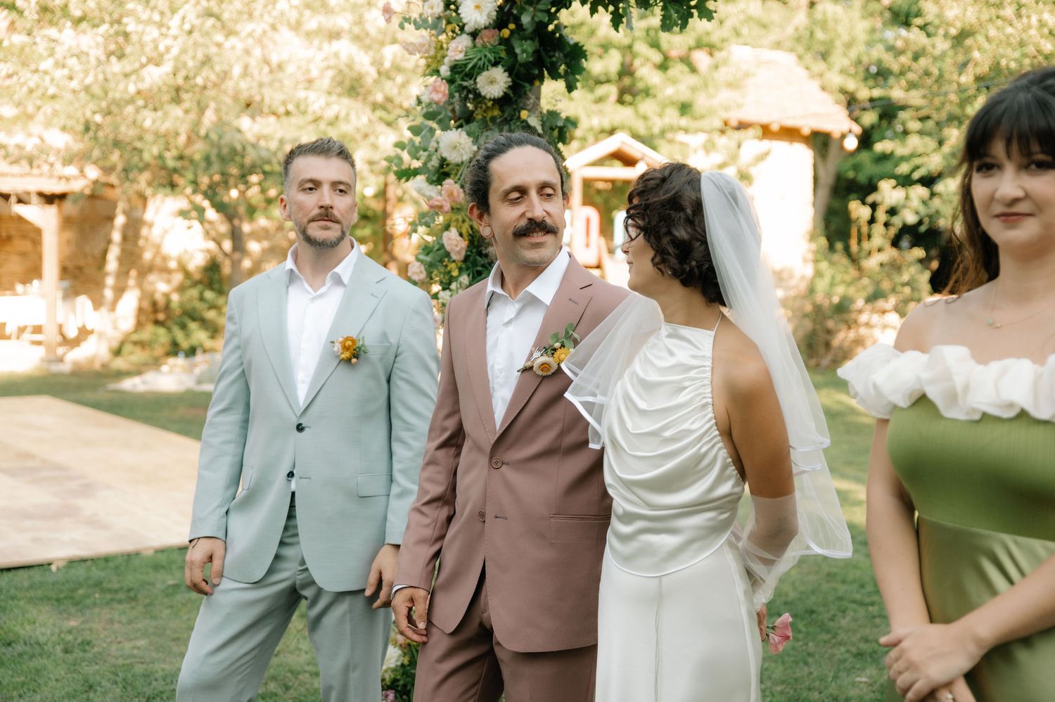 Wedding guests in formal attire gather outdoors in a sunny garden setting during a daytime celebration.