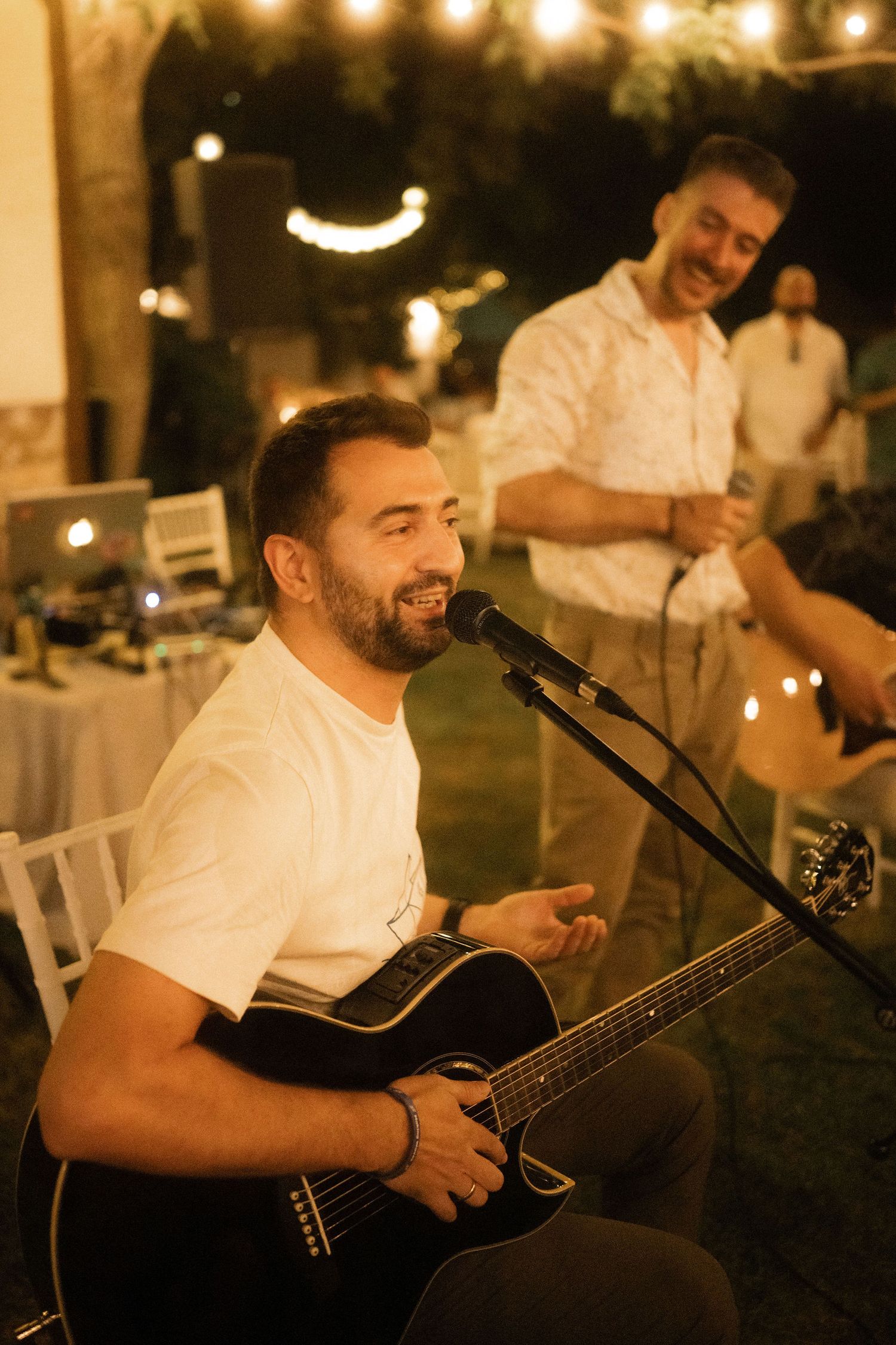 Musician in white shirt plays acoustic guitar and sings at outdoor evening event with string lights overhead.