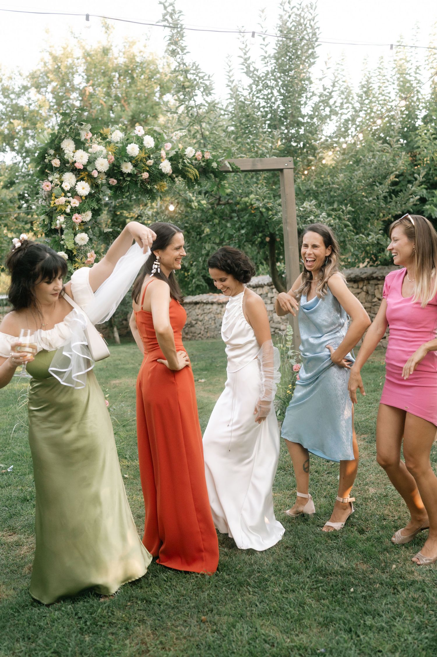 A group in colorful dresses dances joyfully together on a grassy lawn during a summer celebration.