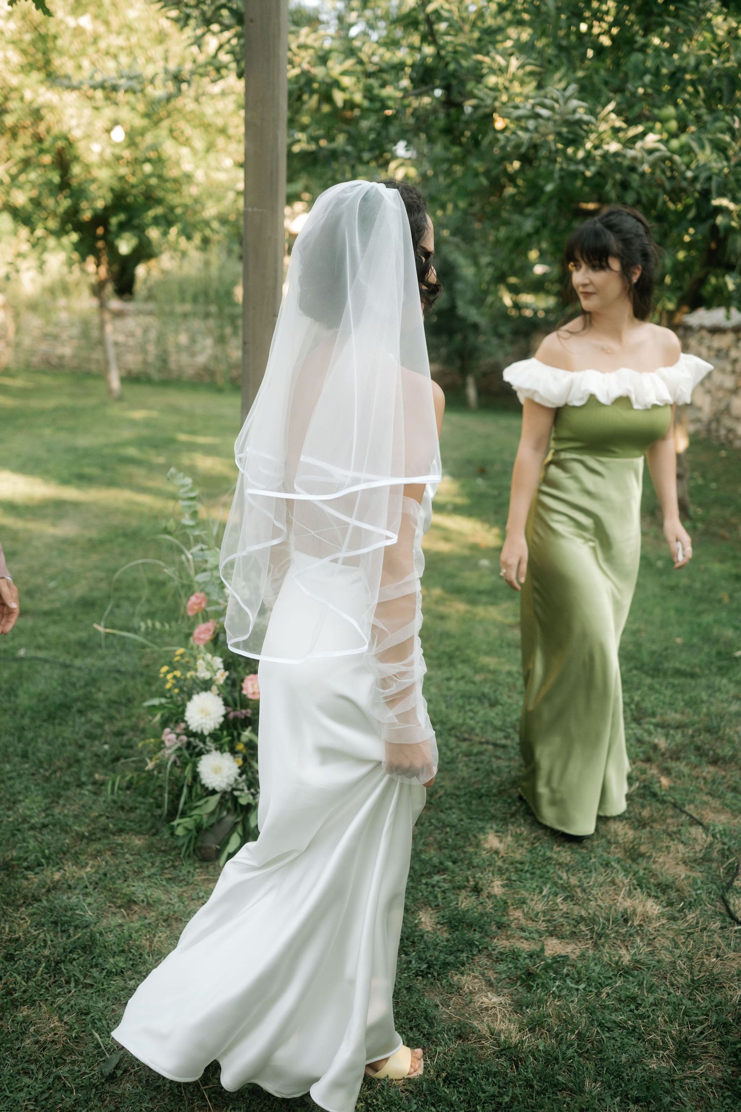 A wedding party walks through a sunlit garden with the bride in white and bridesmaids in sage green dresses.