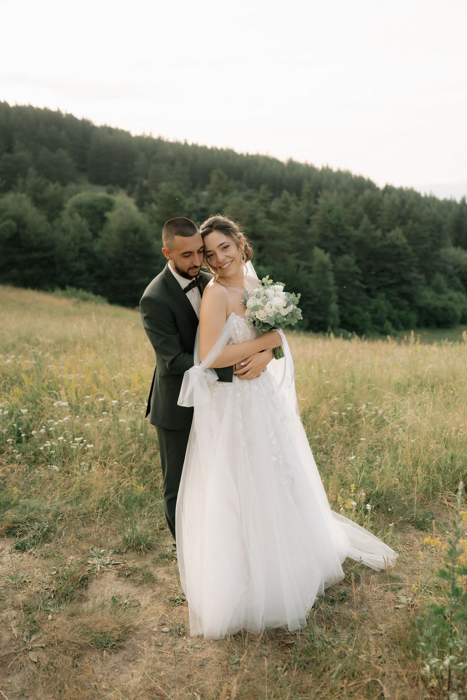 A romantic wedding photo of a couple embracing in a meadow with evergreen forest in the background.