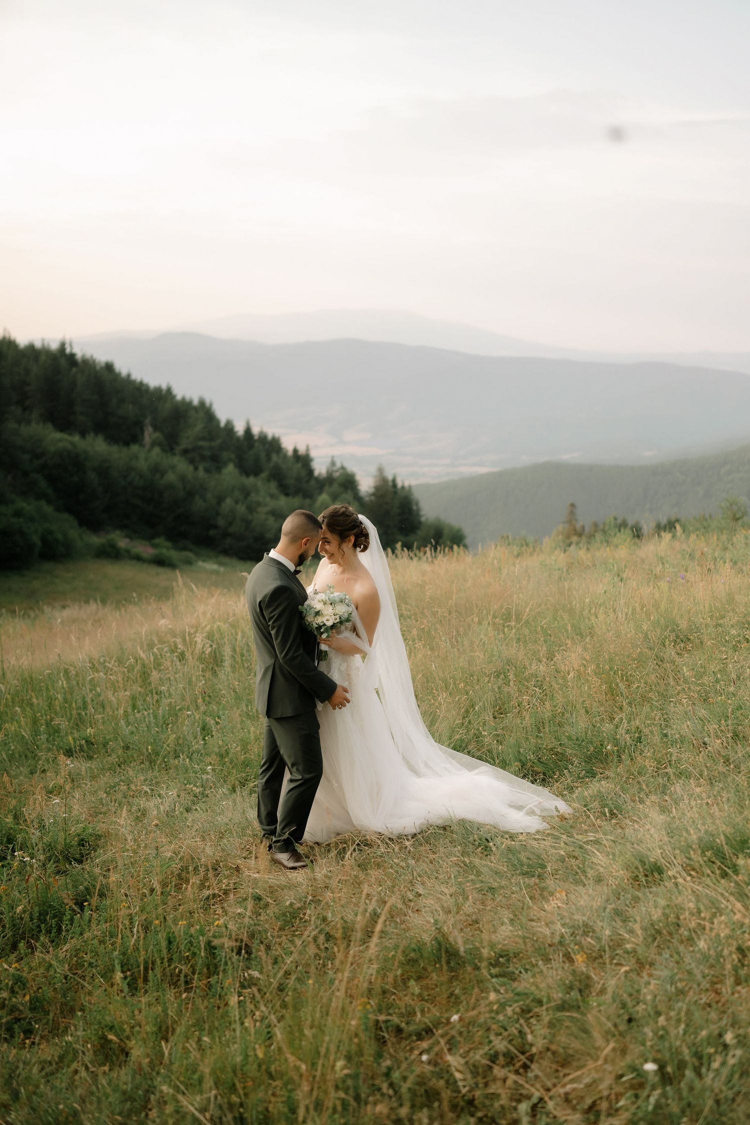 A couple shares an intimate moment in their wedding attire on a misty mountainside meadow with rolling hills in the distance.