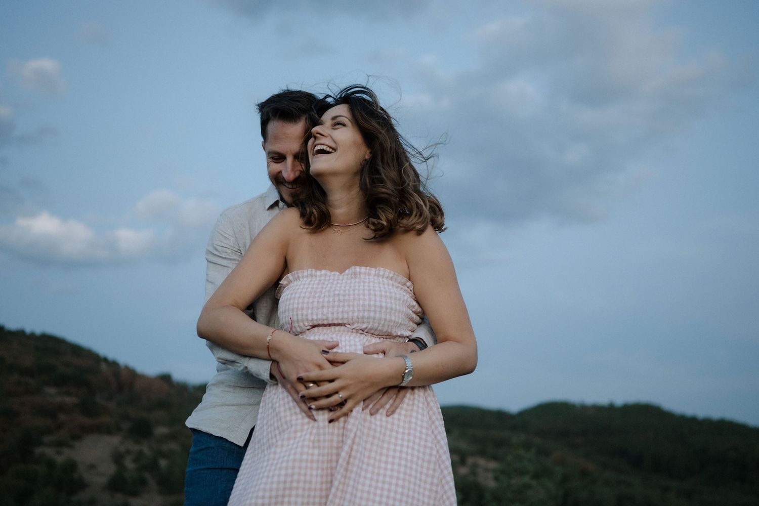 A couple embraces on a mountaintop at dusk wearing light colored clothing against a moody cloudy sky.