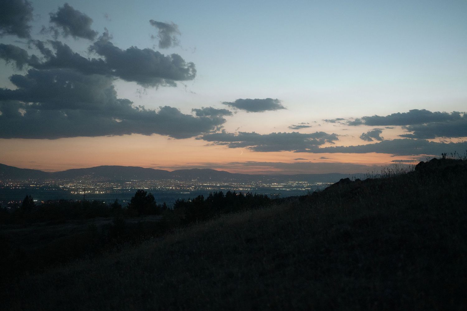A dusky cityscape view from a hilltop shows twinkling lights below as clouds drift across the sunset sky.