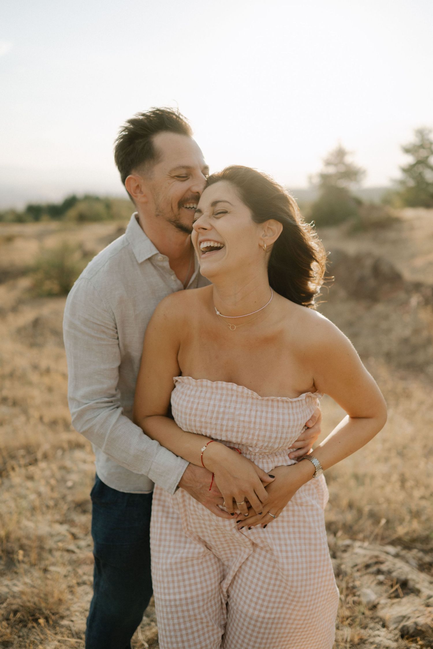 A couple embraces and laughs together in a sunny outdoor setting with golden light and desert terrain.