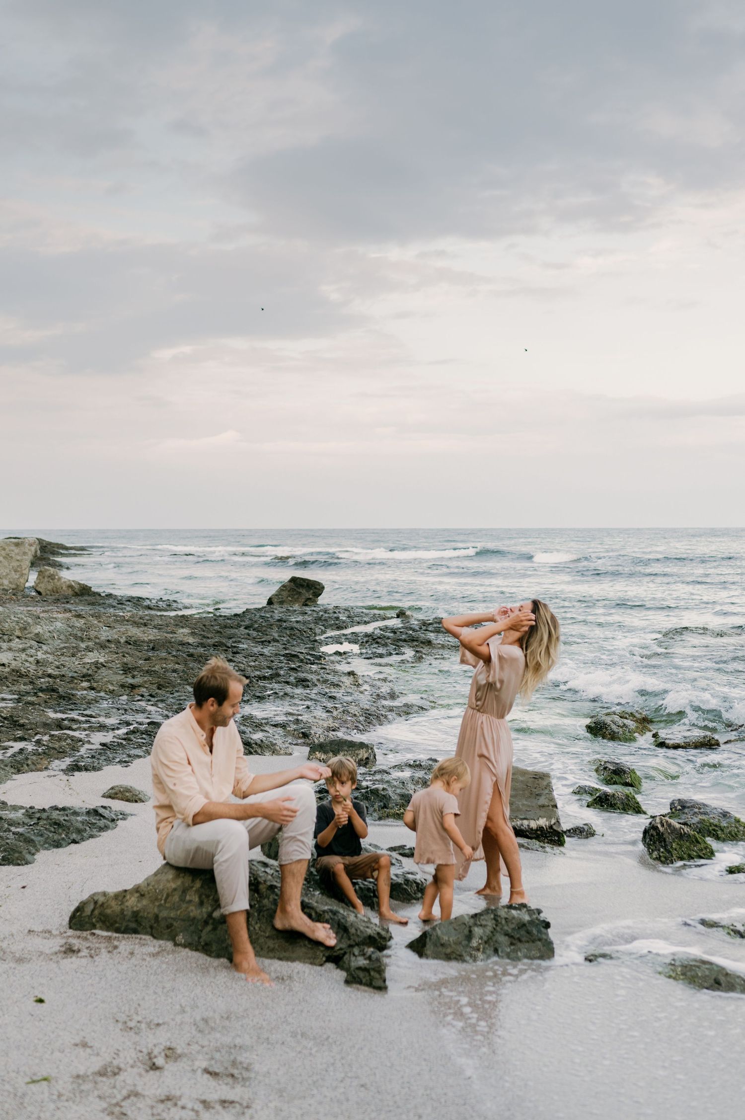 Family of four enjoying a rocky beach at sunset, with waves and cloudy skies in the background.