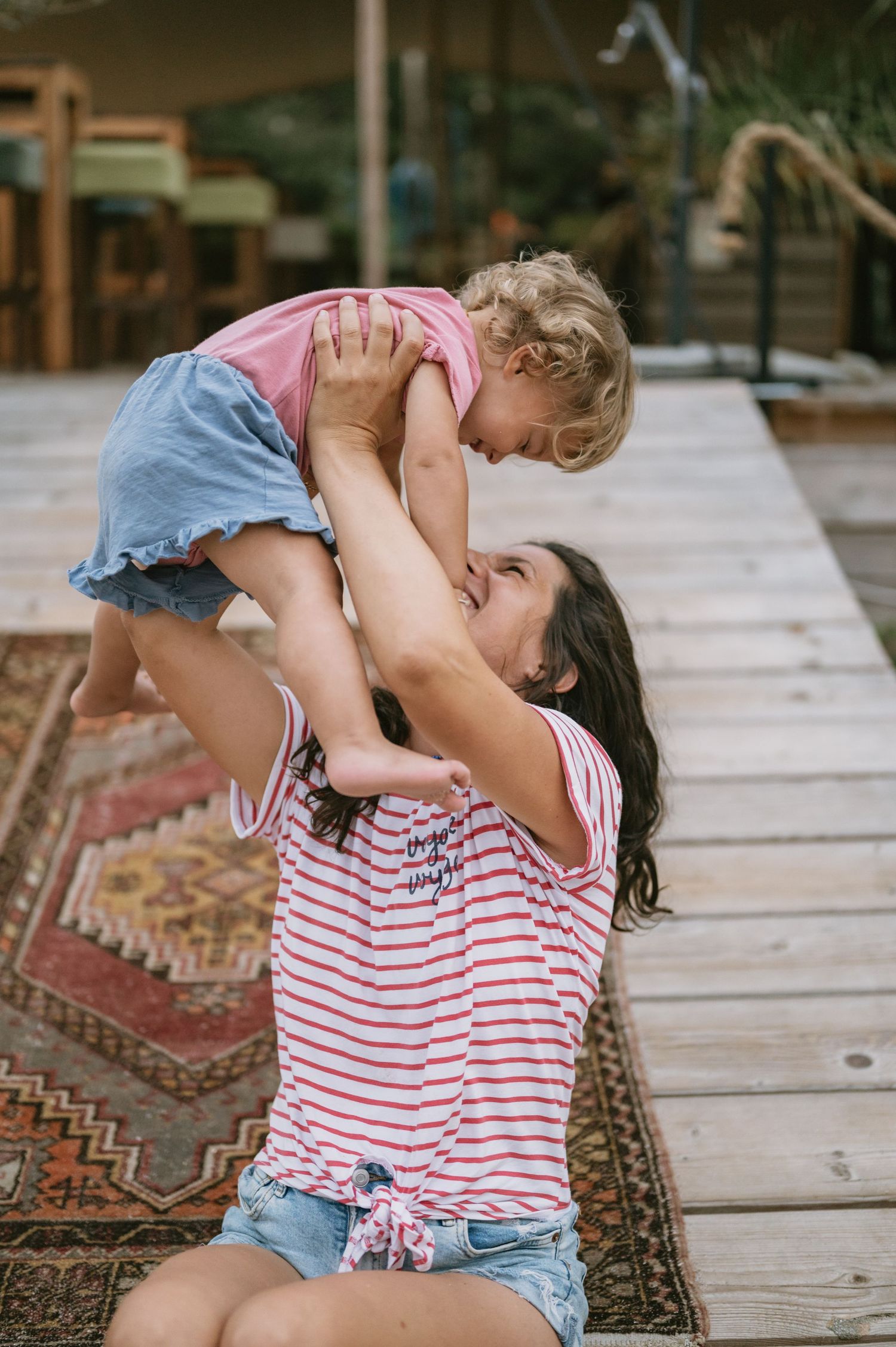 A mother lifts her young child into the air while sitting on a wooden deck, both smiling and sharing a joyful moment.