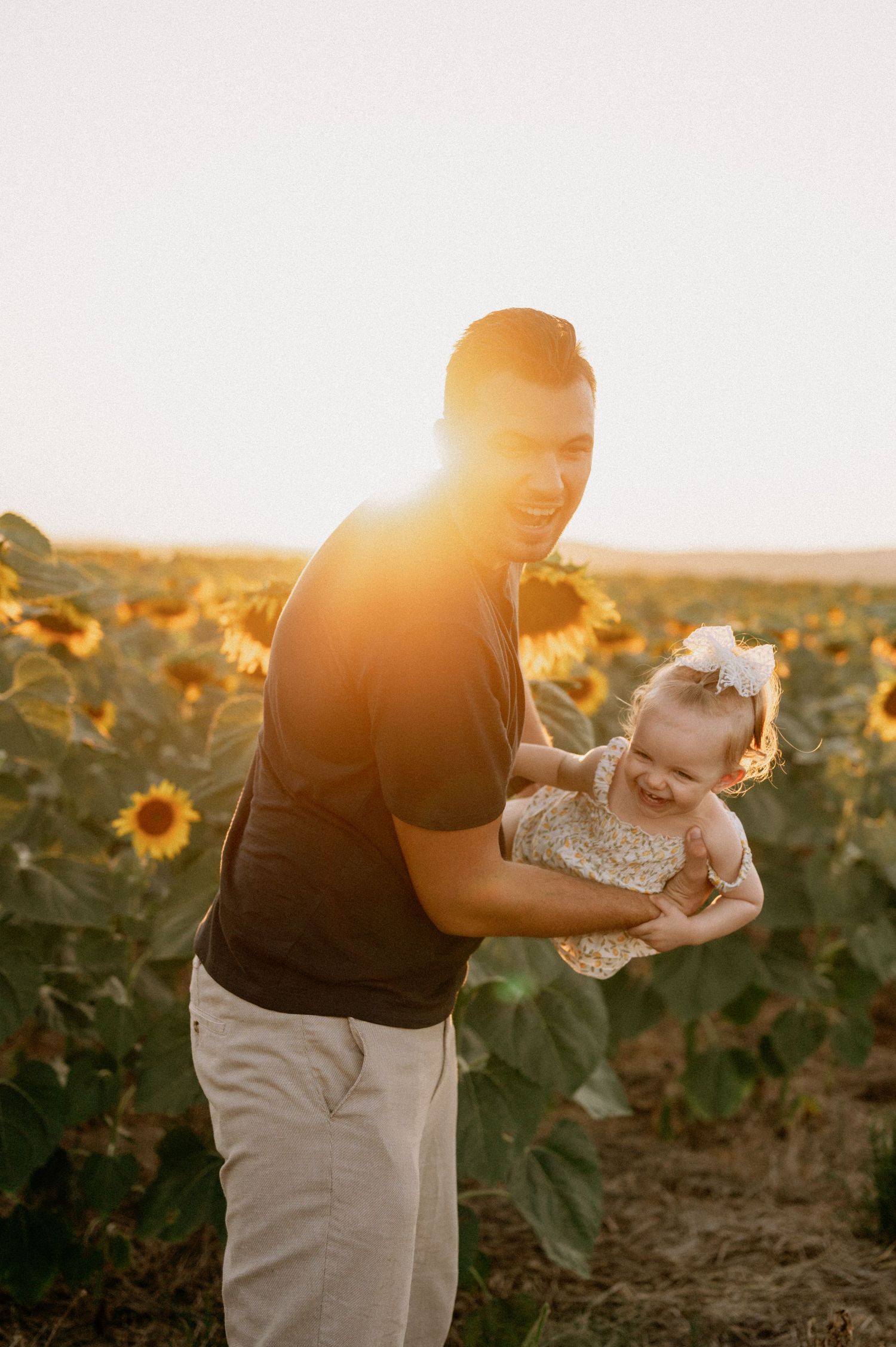 A man holds a smiling baby in a sunflower field at golden hour sunset.
