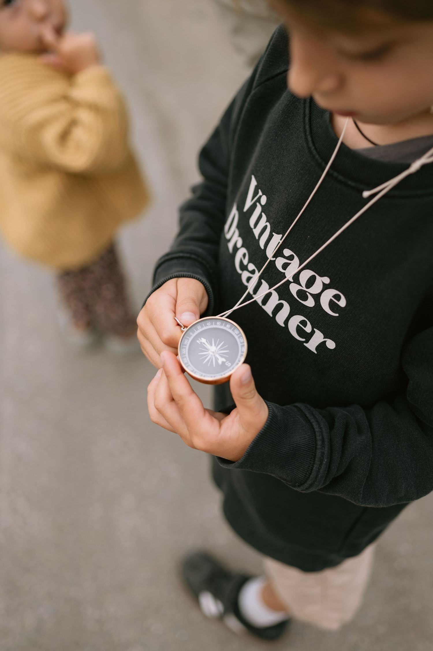 Child in Vintage Dreamer sweatshirt holding a compass necklace, with another child in yellow sweater in background.