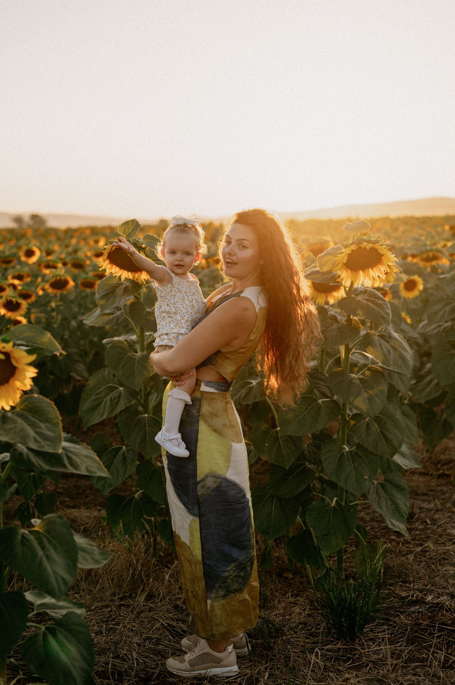 Woman in yellow dress holding toddler in sunflower field at golden hour sunset.