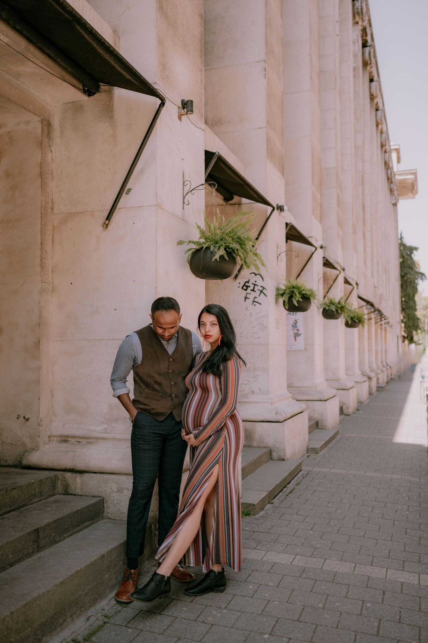 Couple posing by grand stone columns, woman in colorful striped dress, man in vest, urban architecture backdrop.