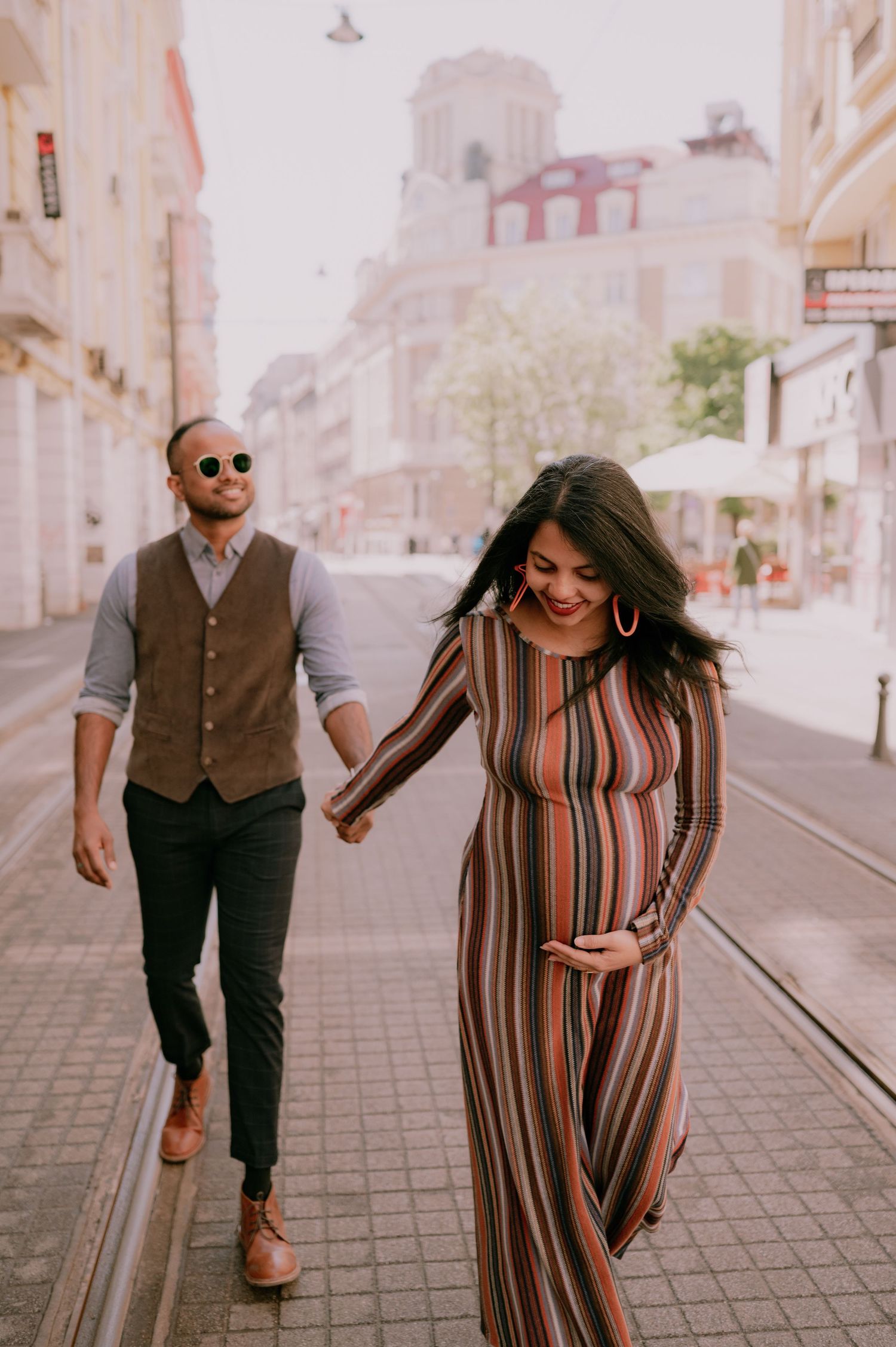 Pregnant woman in striped maxi dress holds hands with stylish man in vest on a city street.