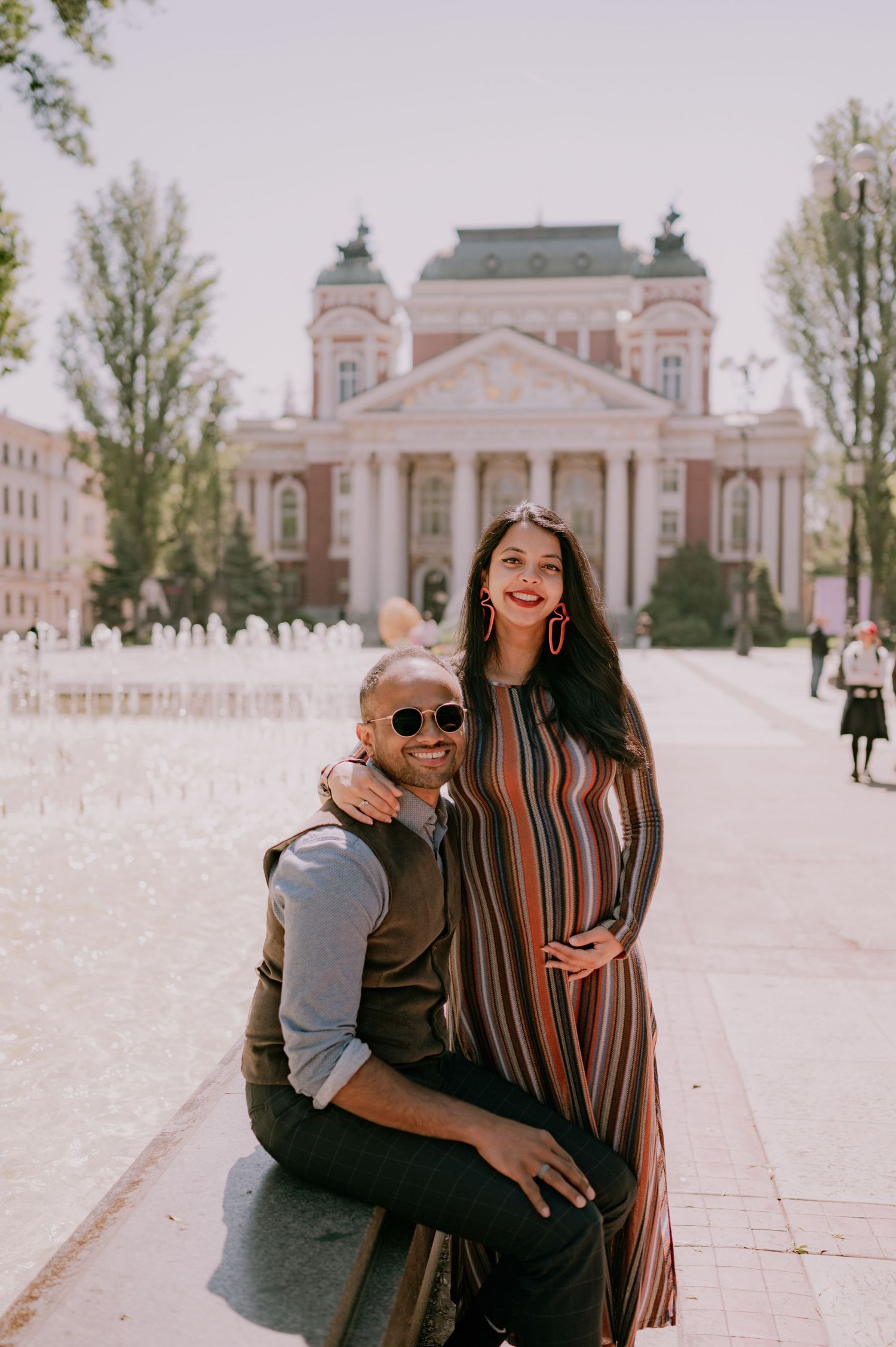 Couple posing in front of a grand neoclassical building with fountains, woman in striped dress, man in sunglasses.