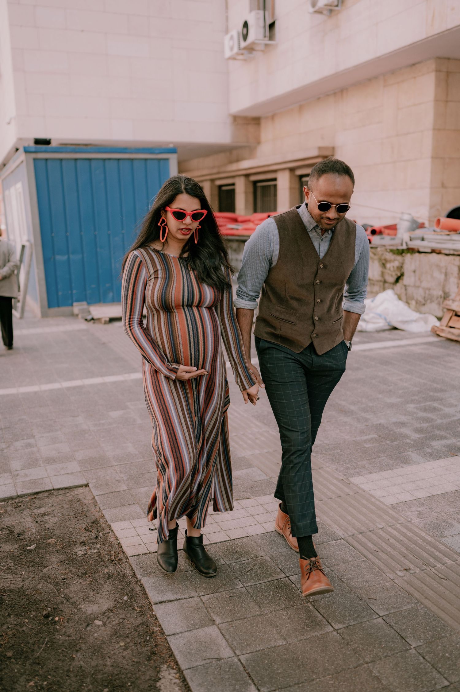 Pregnant woman in striped maxi dress walks hand-in-hand with stylish man in vest and sunglasses on urban sidewalk.