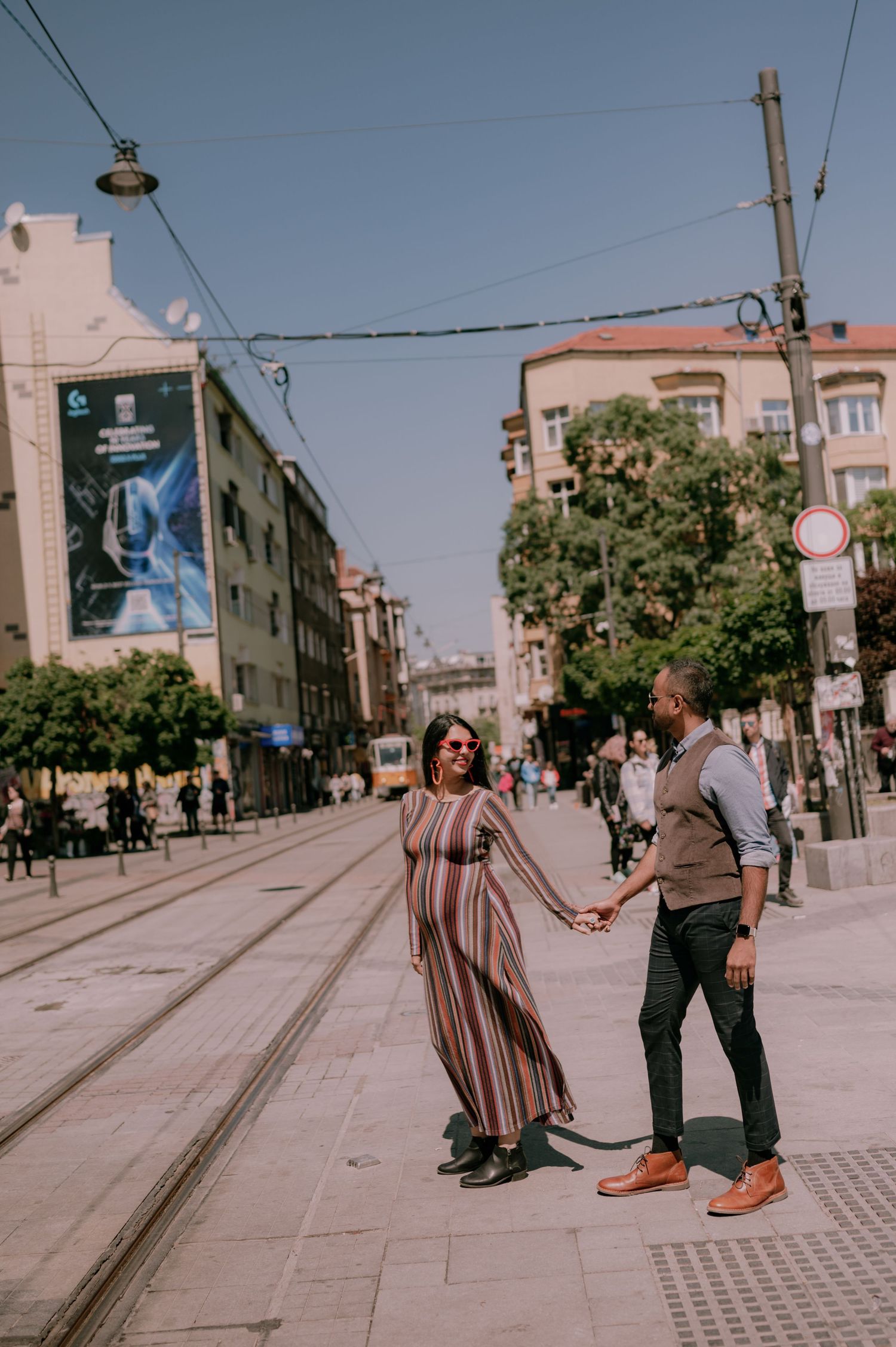 Couple holding hands on a sunny urban street with tram tracks, colorful striped dress, city buildings and trees in background.
