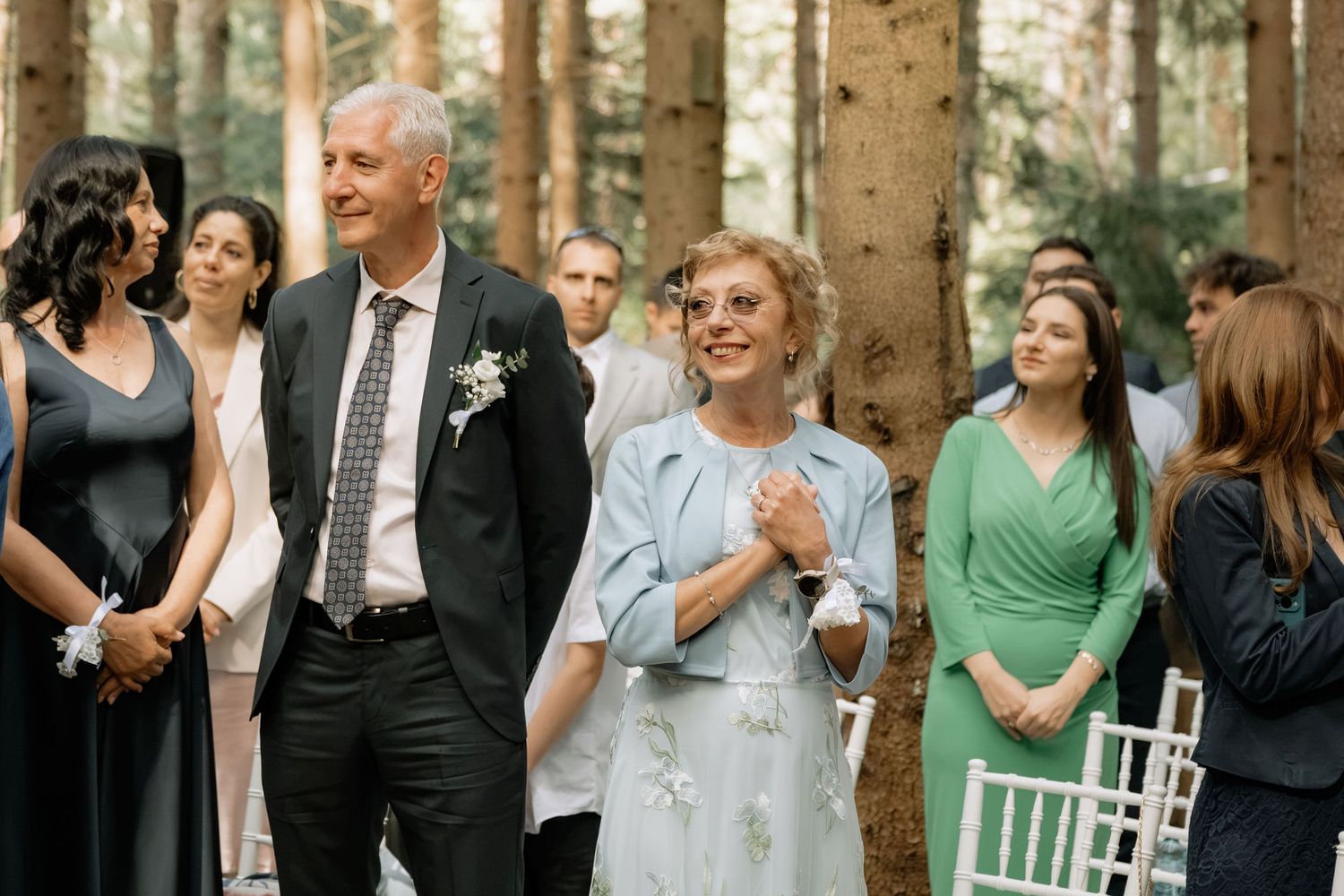 Elegant wedding guests smiling outdoors in a forest ceremony, man in suit with boutonniere, woman in light blue dress.