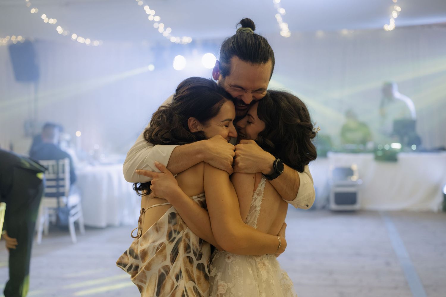 Group hug at a wedding reception with string lights, man and two women embracing on the dance floor.