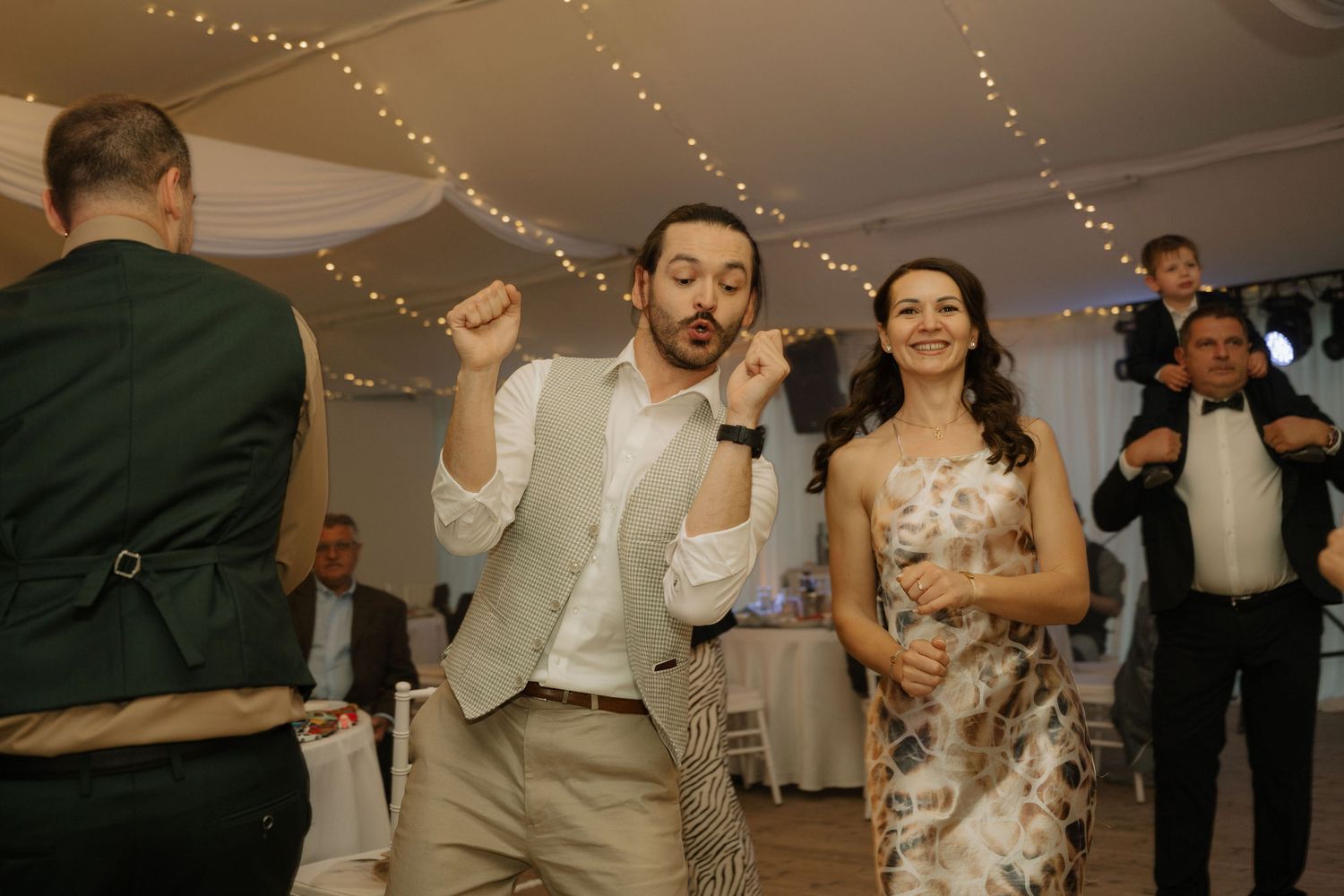 Guests dancing joyfully at a wedding reception with fairy lights and elegant decor in the background.