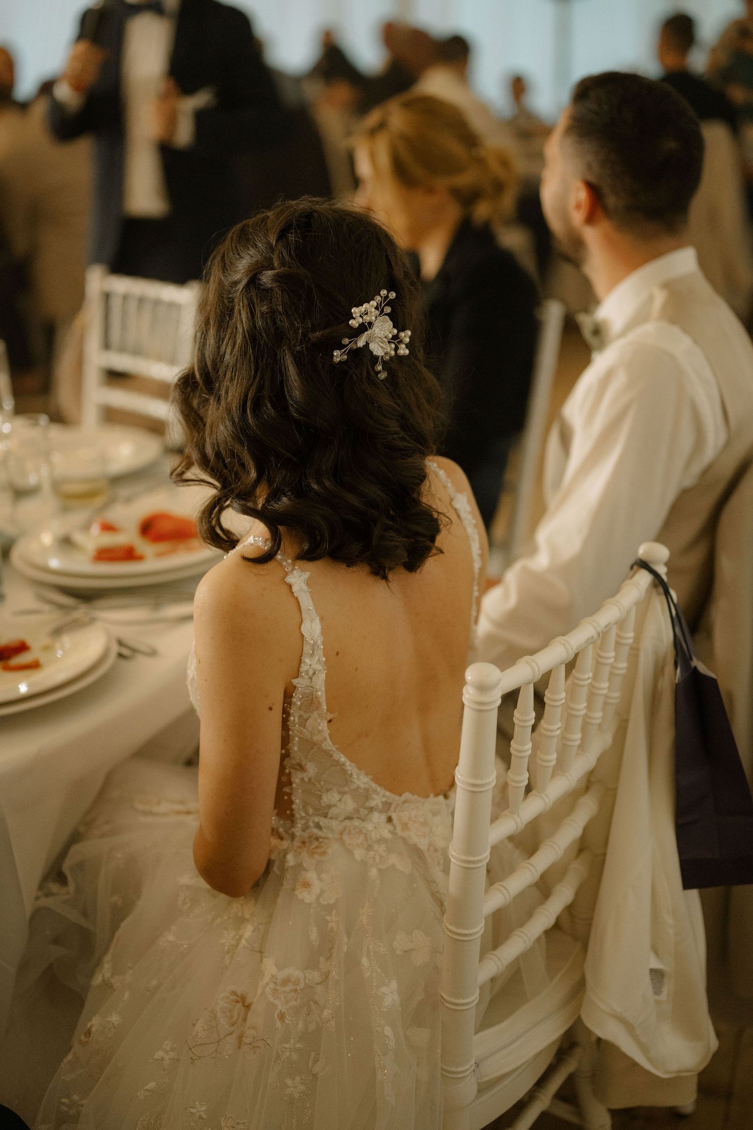 Bride in lace wedding gown with floral hair accessory seated at reception table beside groom in white suit.