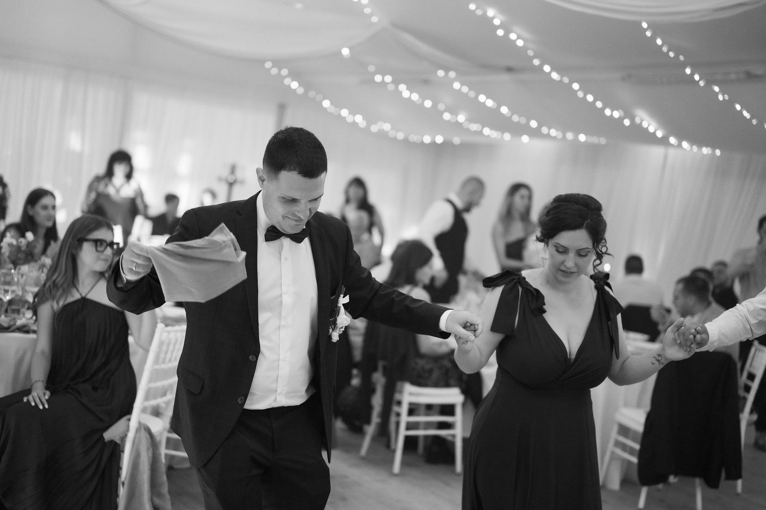 Black and white photo of elegantly dressed guests dancing at a wedding reception in a decorated marquee.
