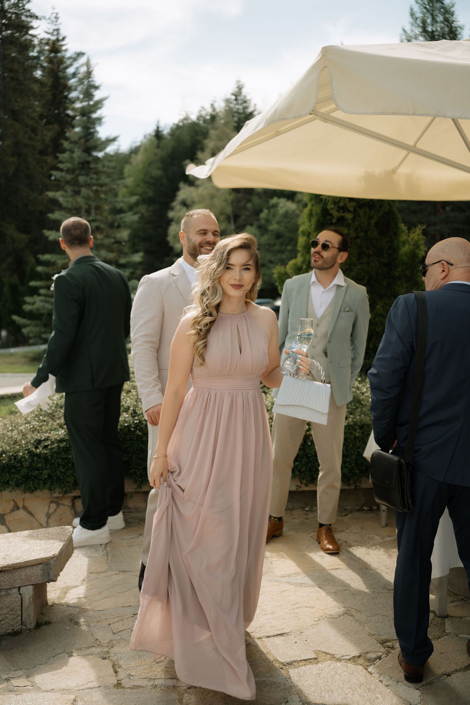 Elegant bridesmaid in blush pink maxi dress walks outdoors at wedding venue with guests in formal attire.