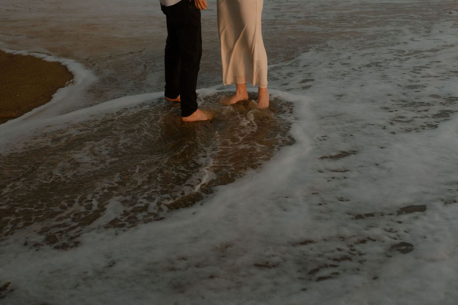 Two barefoot people standing together at the ocean shoreline with gentle waves washing over their feet at sunset.
