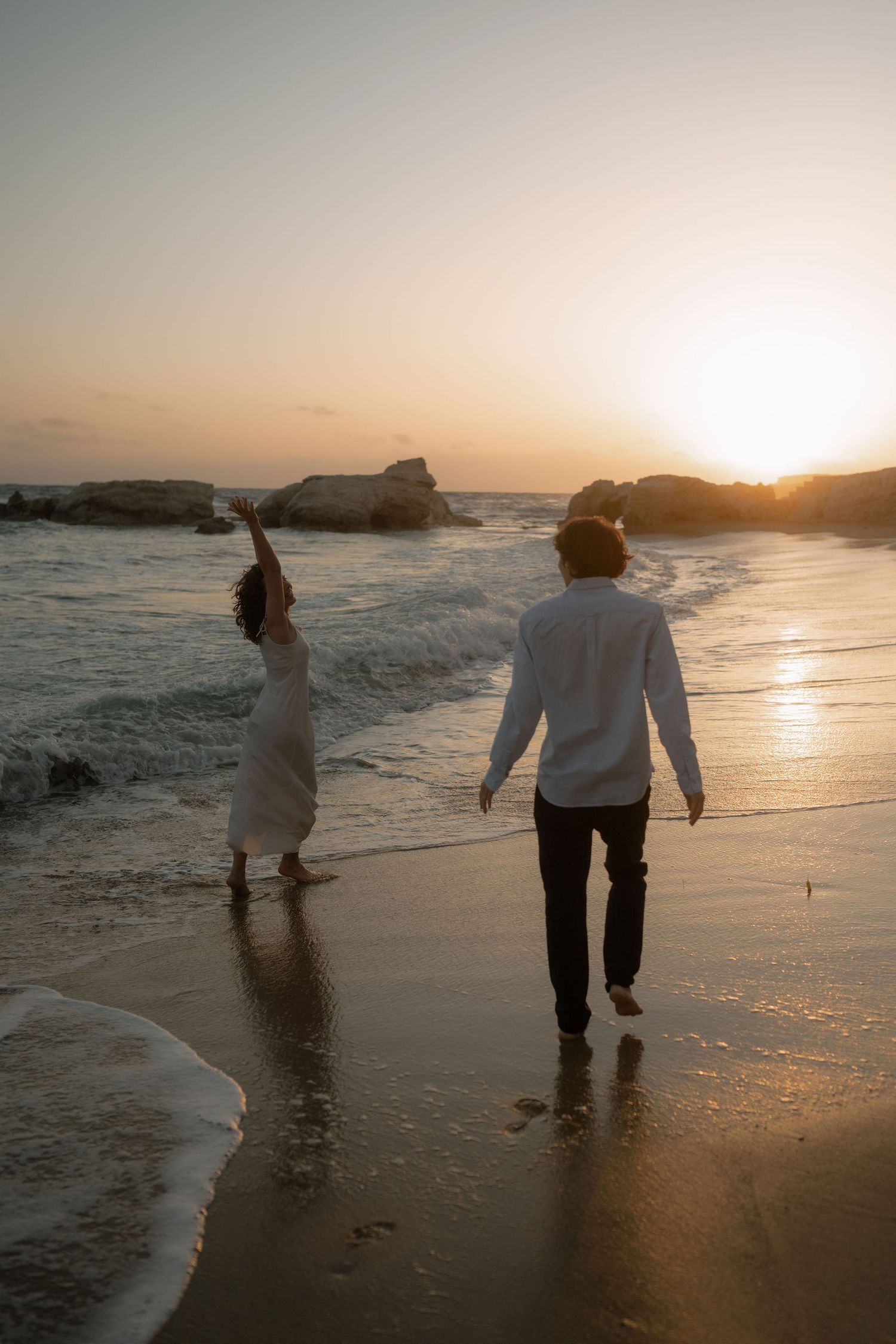 Couple walking on a sandy beach at sunset, woman in white dress raising her arm, man in white shirt and black pants.