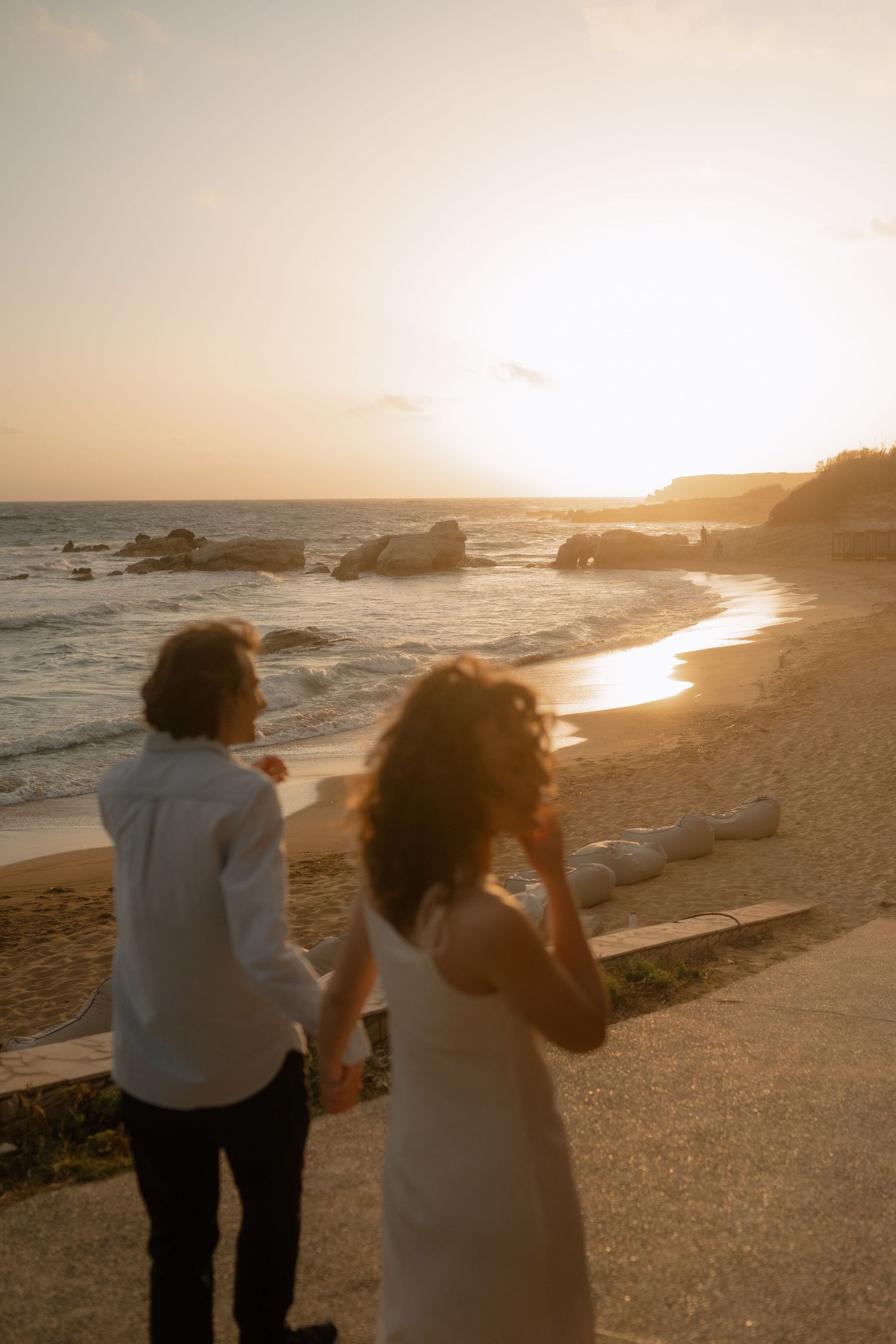 Couple in white attire standing on a sandy beach at golden sunset, waves and rocks visible in warm evening light.