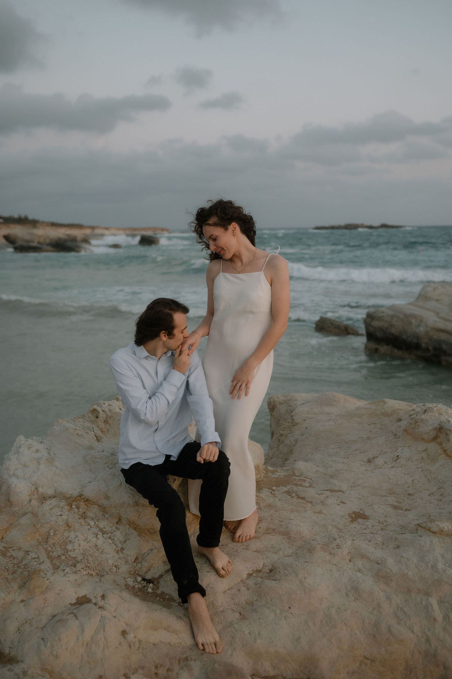 Couple sharing a romantic moment on rocky beach at dusk, woman in cream dress, man in blue shirt, ocean waves behind.