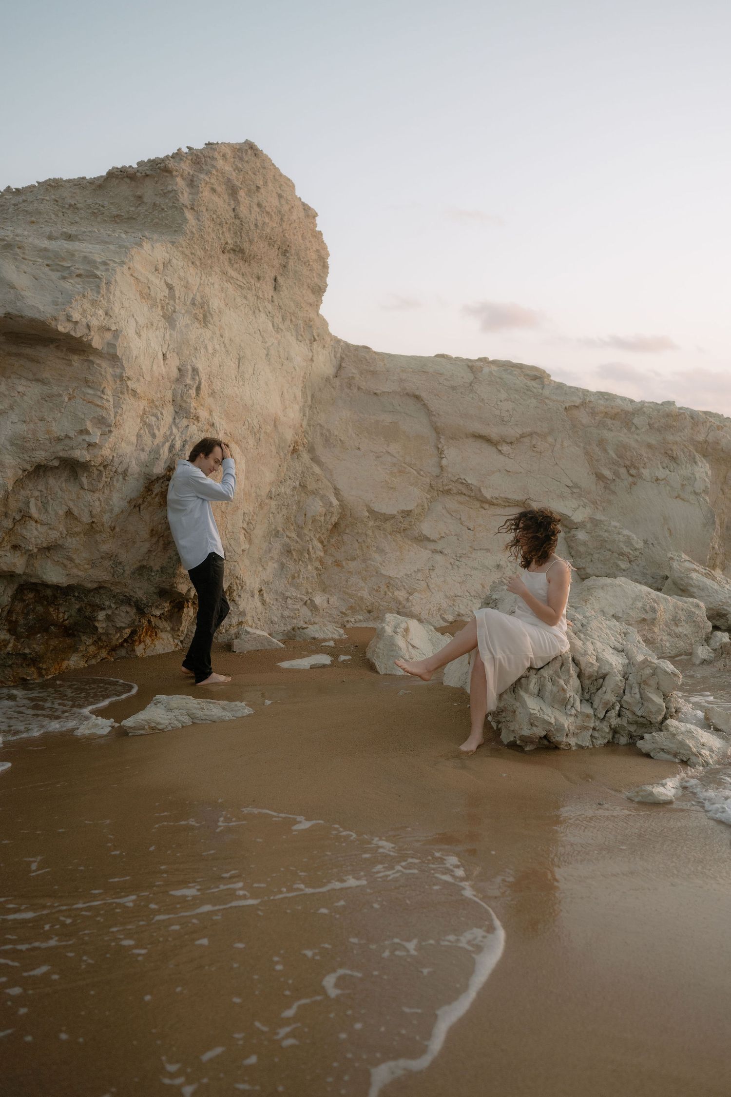 Couple near rocky cliffs on sandy beach at dusk, woman in pink dress seated on rock, man in white shirt standing.