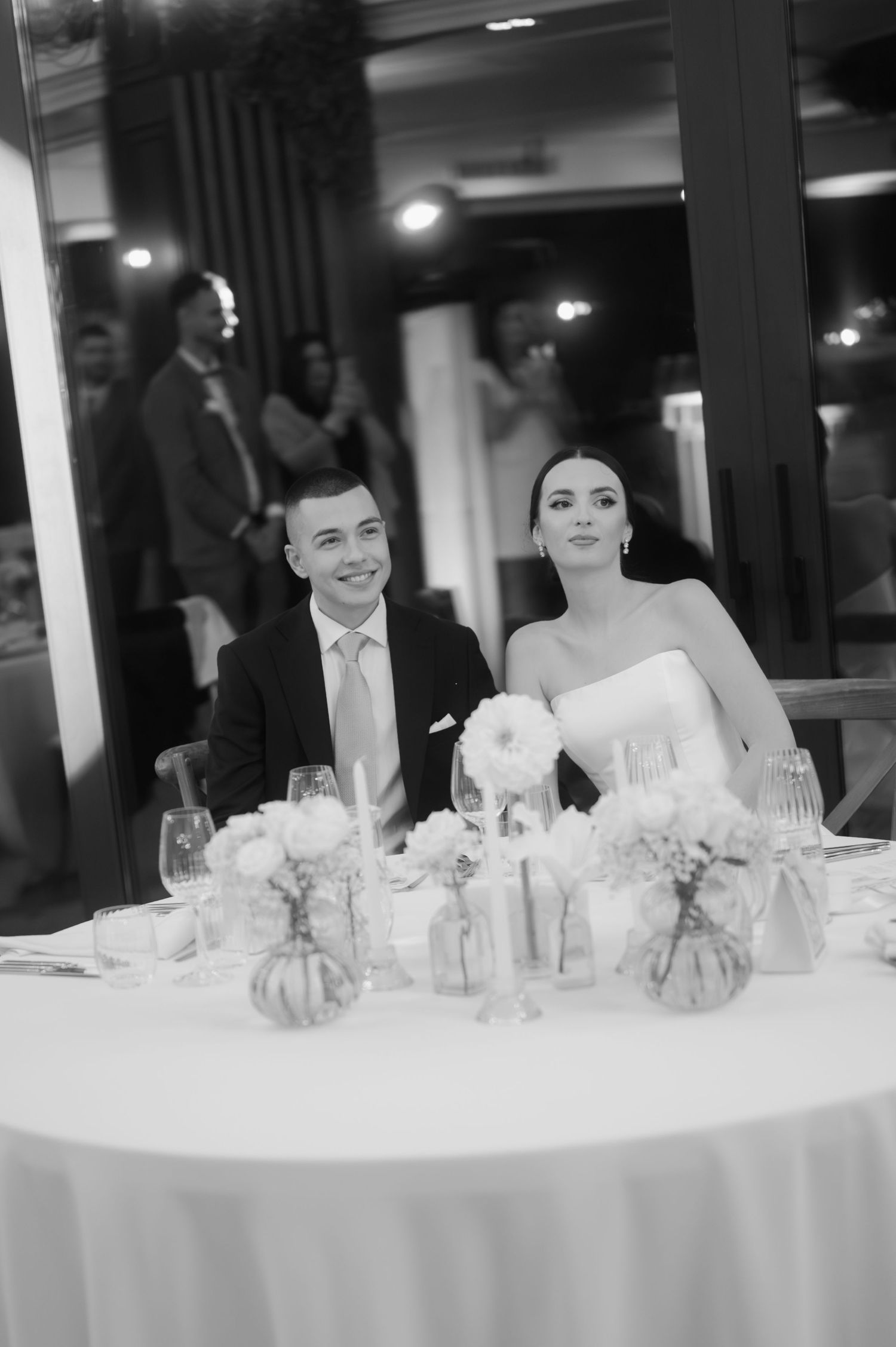 A black and white wedding reception photo showing a couple seated at an elegantly decorated table with floral centerpieces.