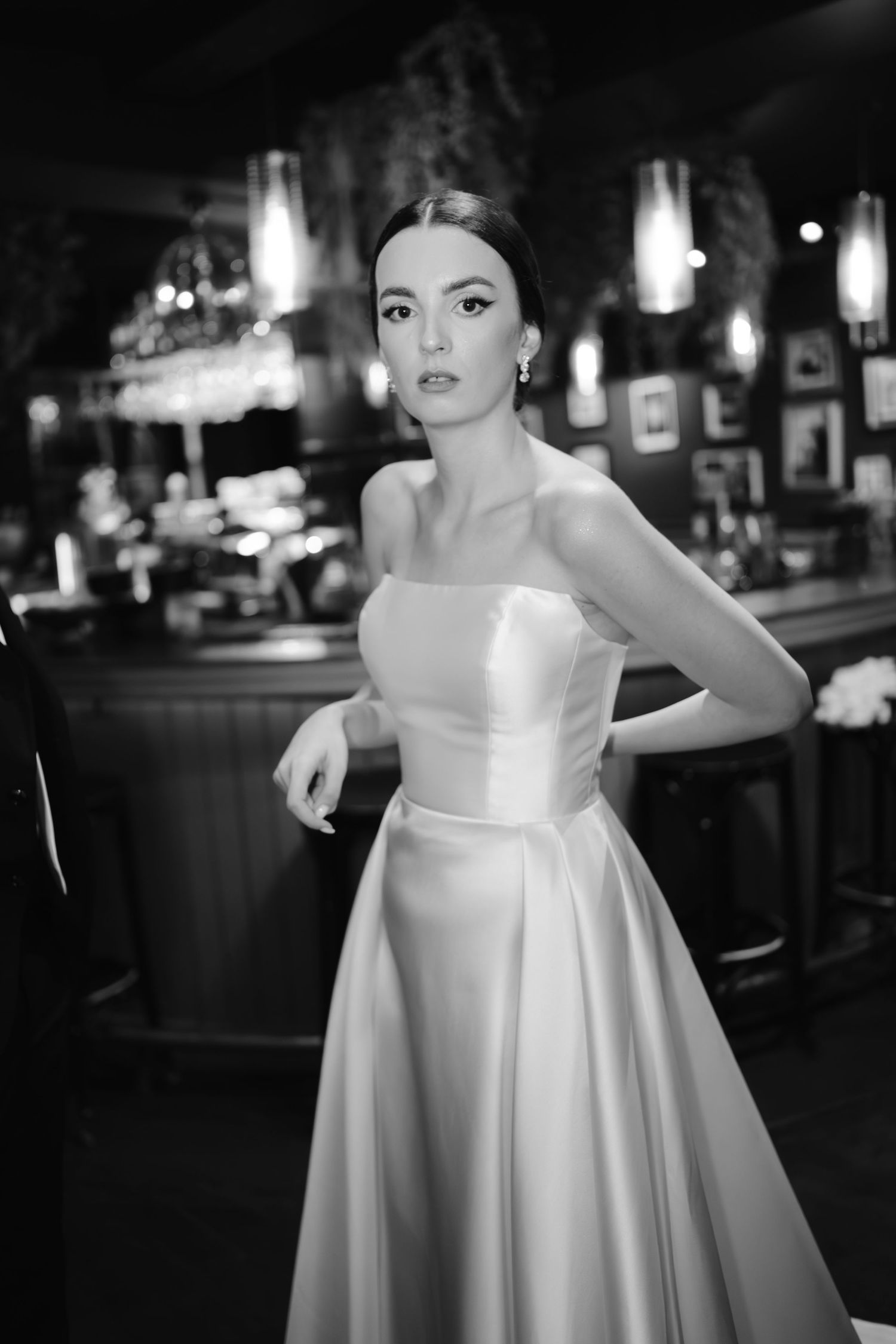 Elegant black and white portrait of someone in a strapless satin wedding gown at a dimly lit bar setting.