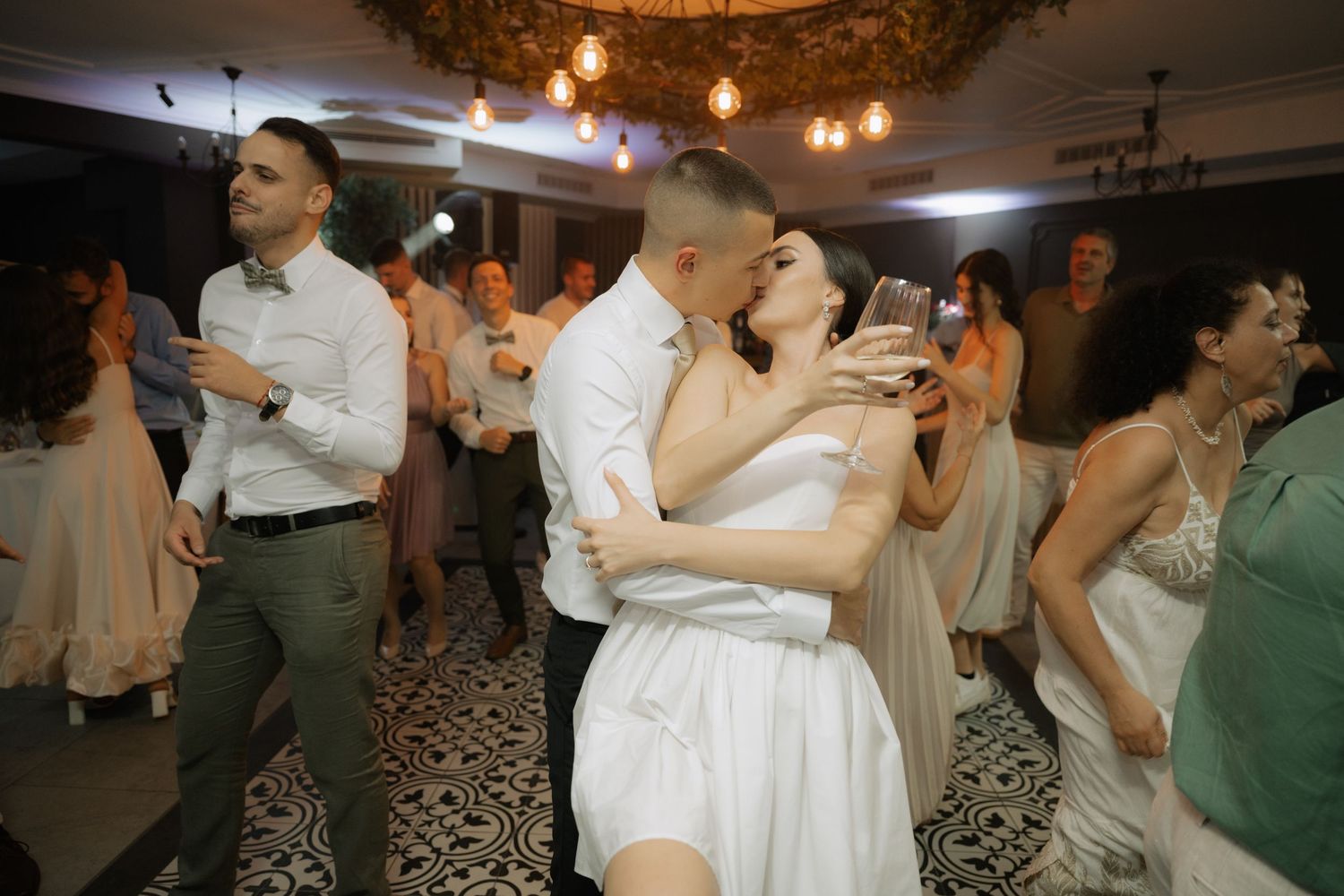 Couples dancing together at a lively indoor wedding reception with string lights overhead.