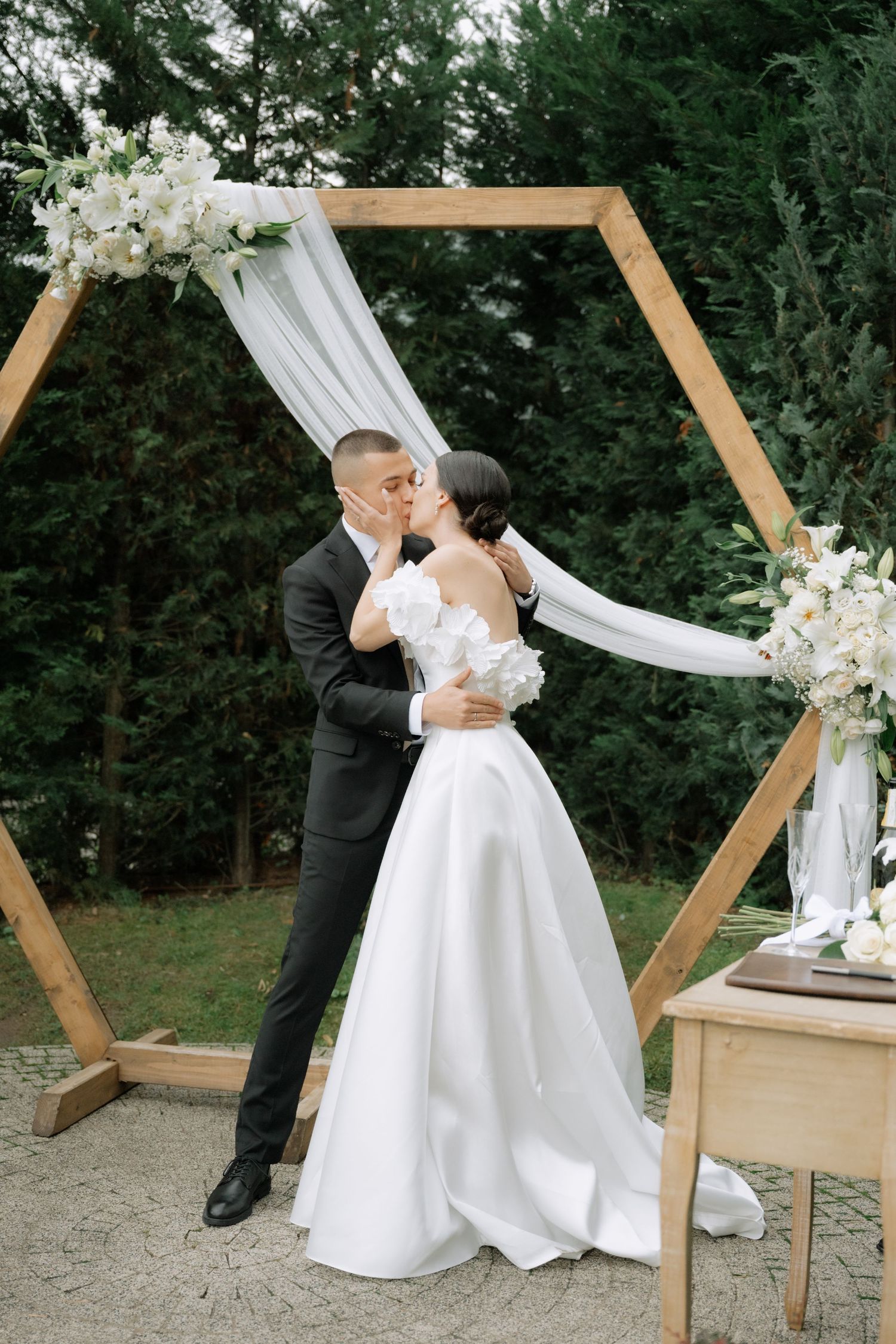 A romantic wedding moment under a hexagonal wooden arch decorated with white flowers and flowing fabric in an outdoor garden setting.