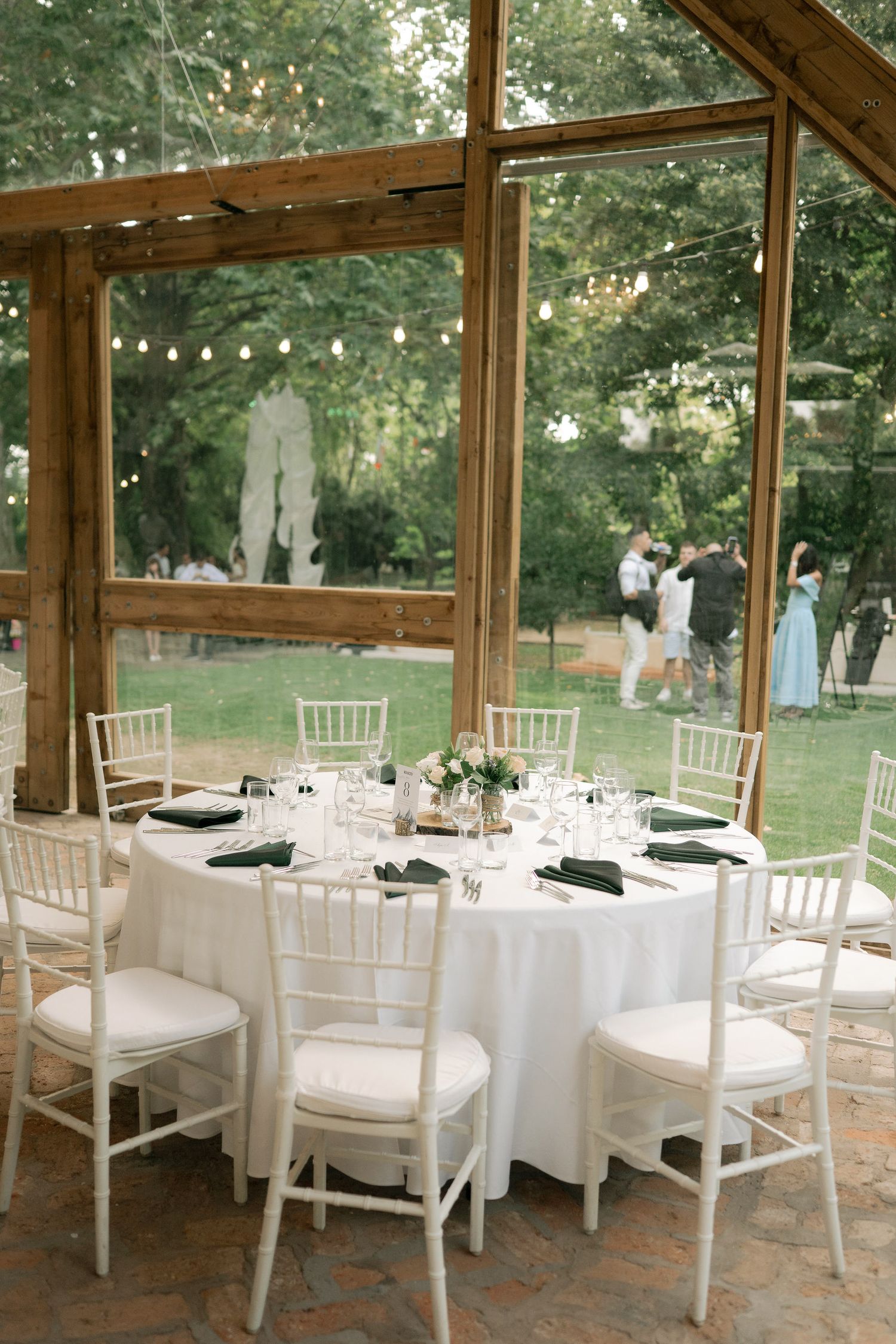 Elegant outdoor wedding reception setup with white tables and chairs under a rustic wooden pergola in a garden setting.