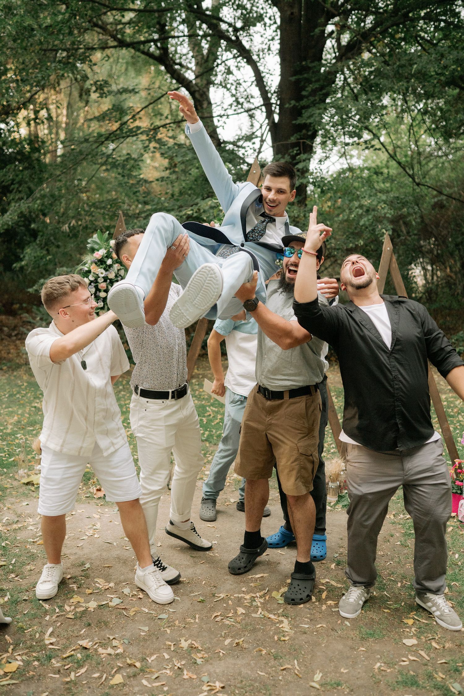 A group of wedding party members jumping and posing playfully in a wooded outdoor setting.