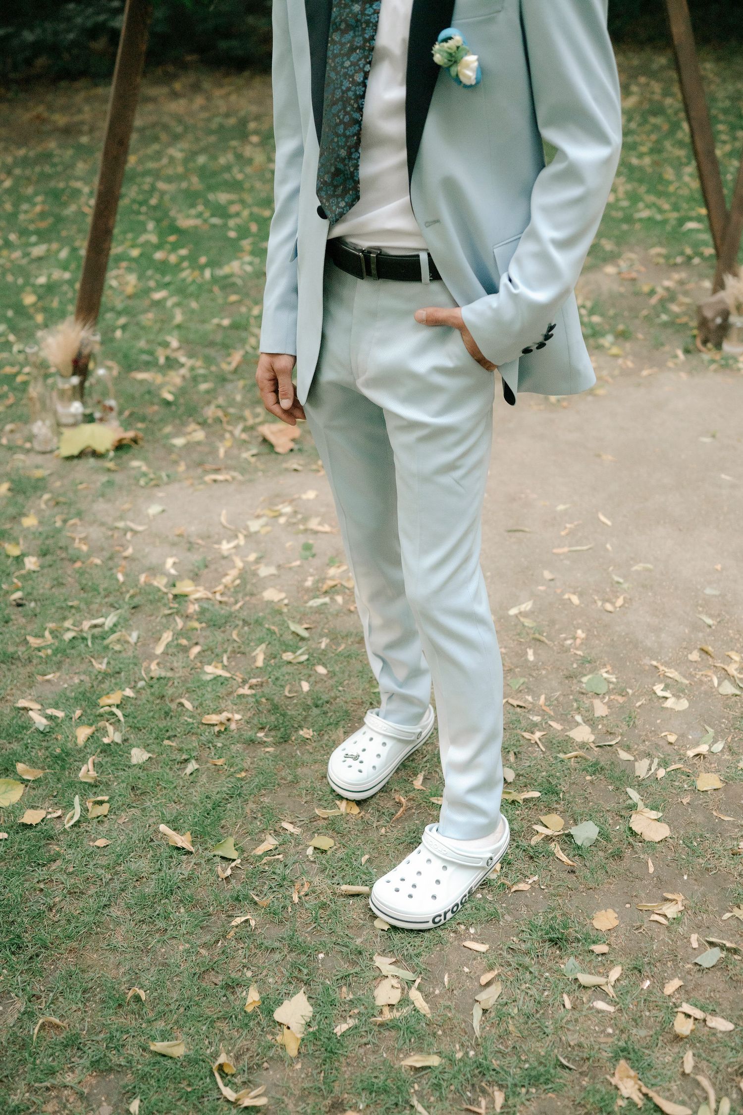 Person wearing light gray suit pants and white sneakers standing on ground covered with fallen autumn leaves.