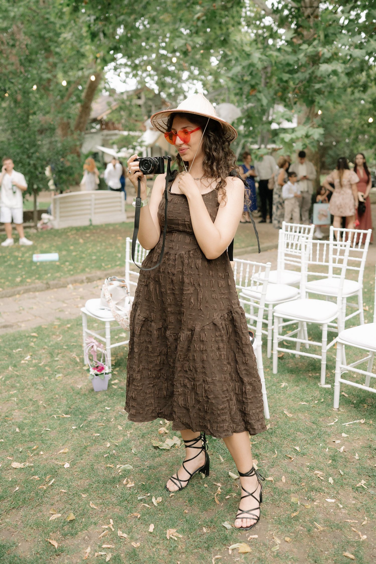 Young child in brown dress and sun hat twirls playfully on outdoor lawn with white chairs in background.
