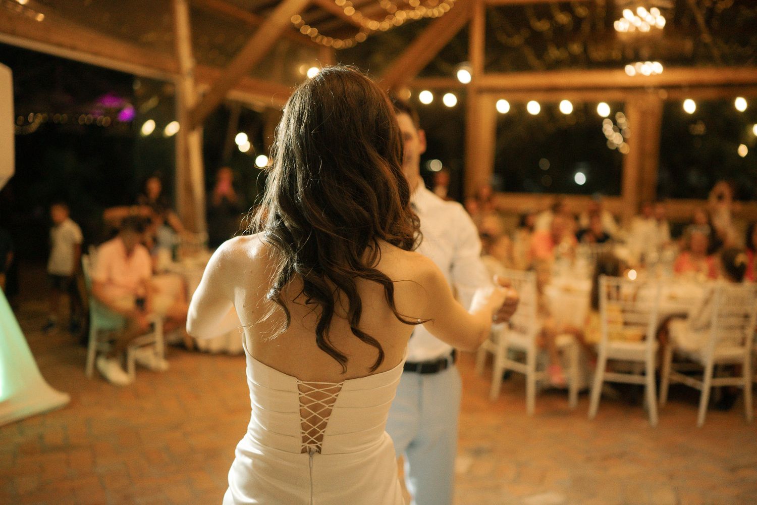 A wedding reception at night with string lights illuminating round tables and guests dancing on an outdoor patio.