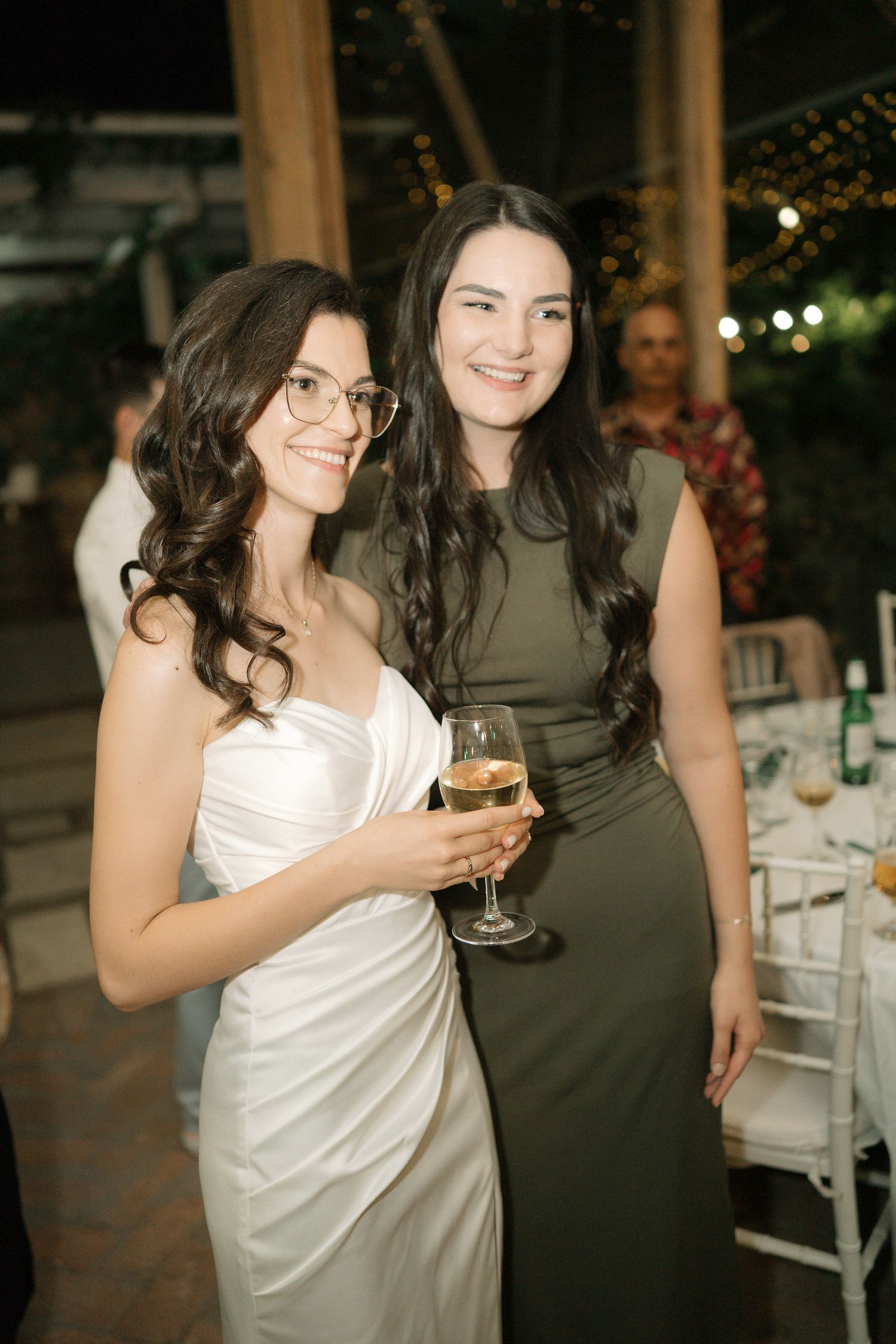 Two friends in contrasting white and dark dresses share laughs and drinks at an outdoor evening event.