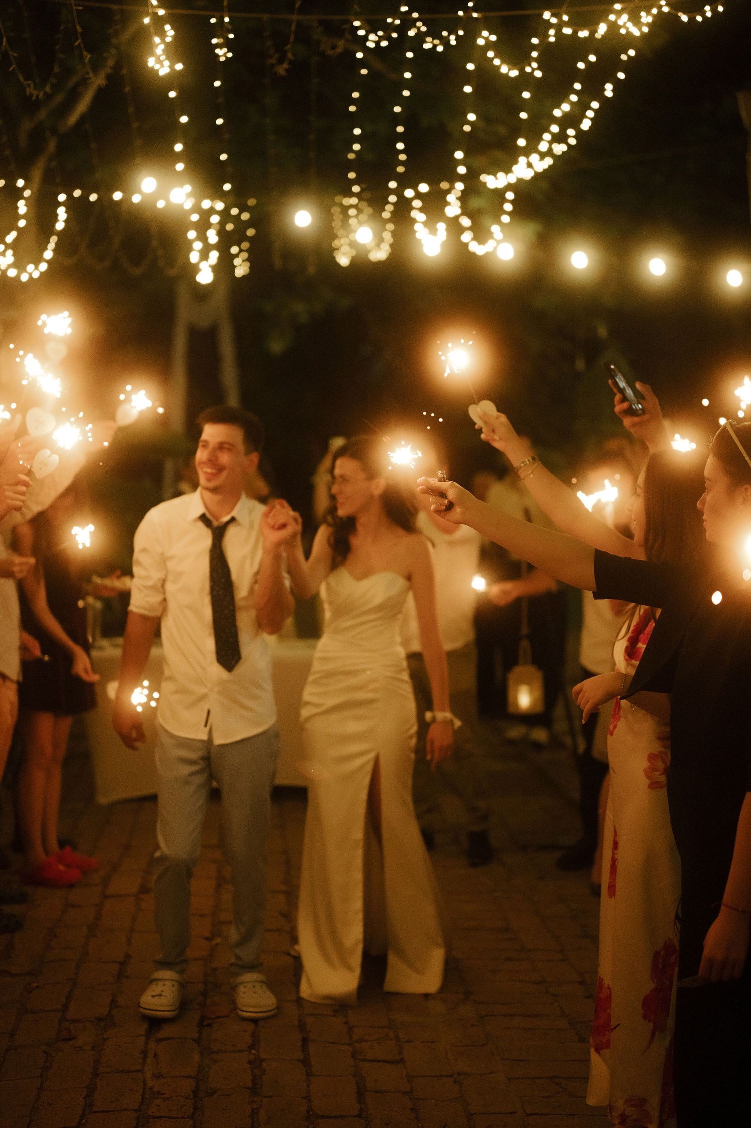 Wedding guests hold sparklers during an evening celebration under twinkling string lights on a brick patio.