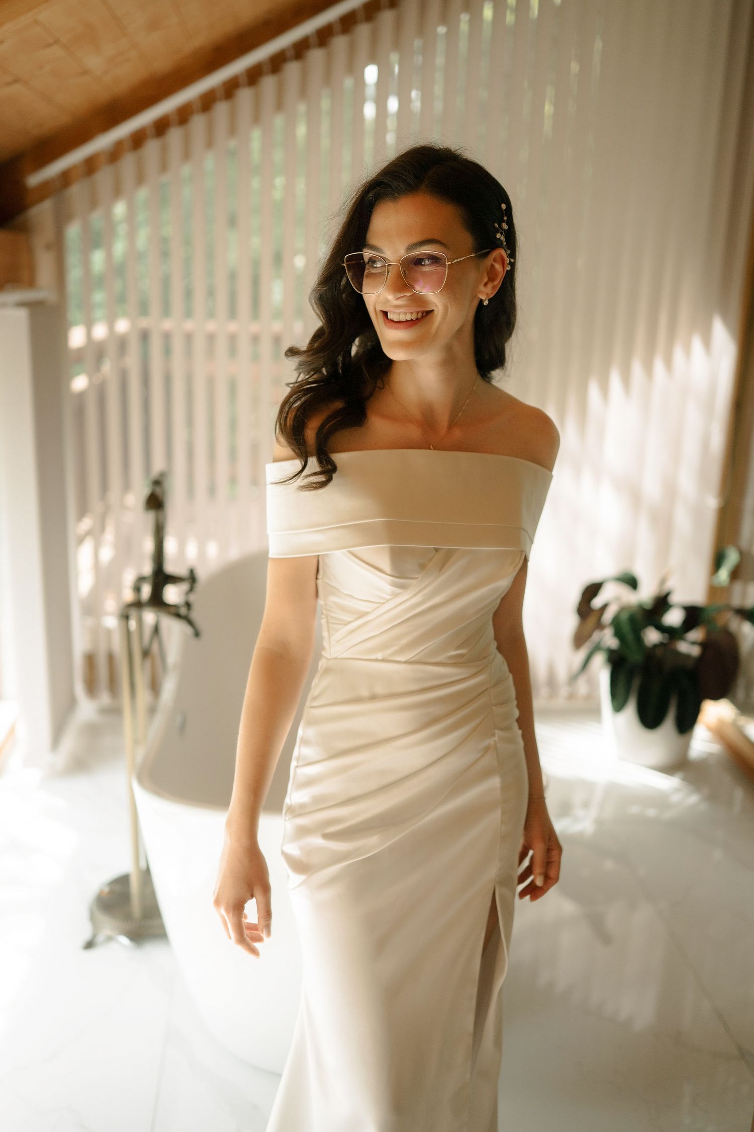 A bride in a sleek off-shoulder white wedding dress and glasses smiles warmly in a sunlit room with white curtains.