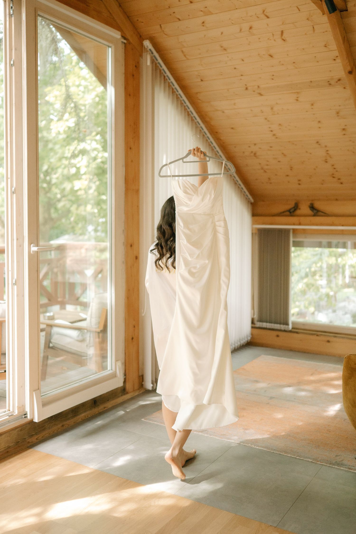 A series of white dresses hanging on wooden hangers in a sunlit room with pine wood walls and windows.