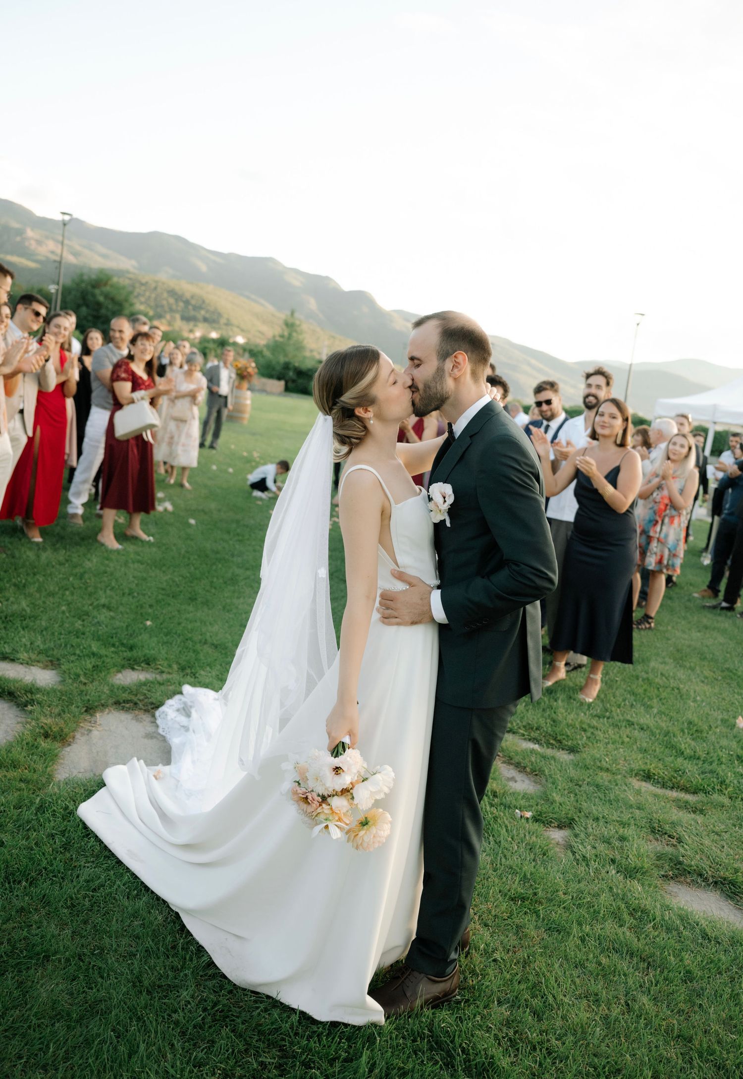Newlyweds share their first dance on a grassy hillside overlooking mountains at an outdoor wedding celebration.