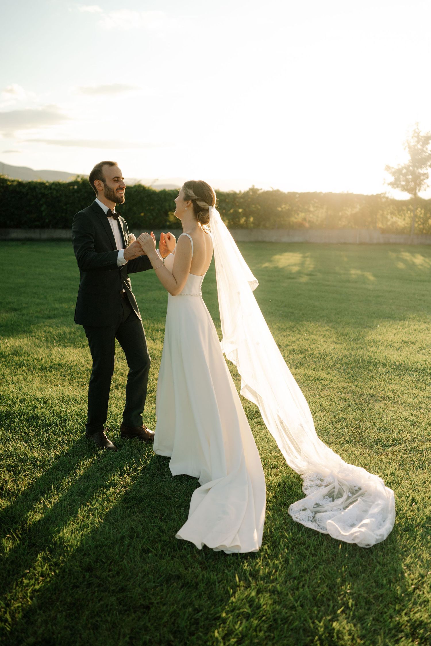 A bride and groom share an intimate moment in a sunlit field during golden hour as her long veil flows in the breeze.